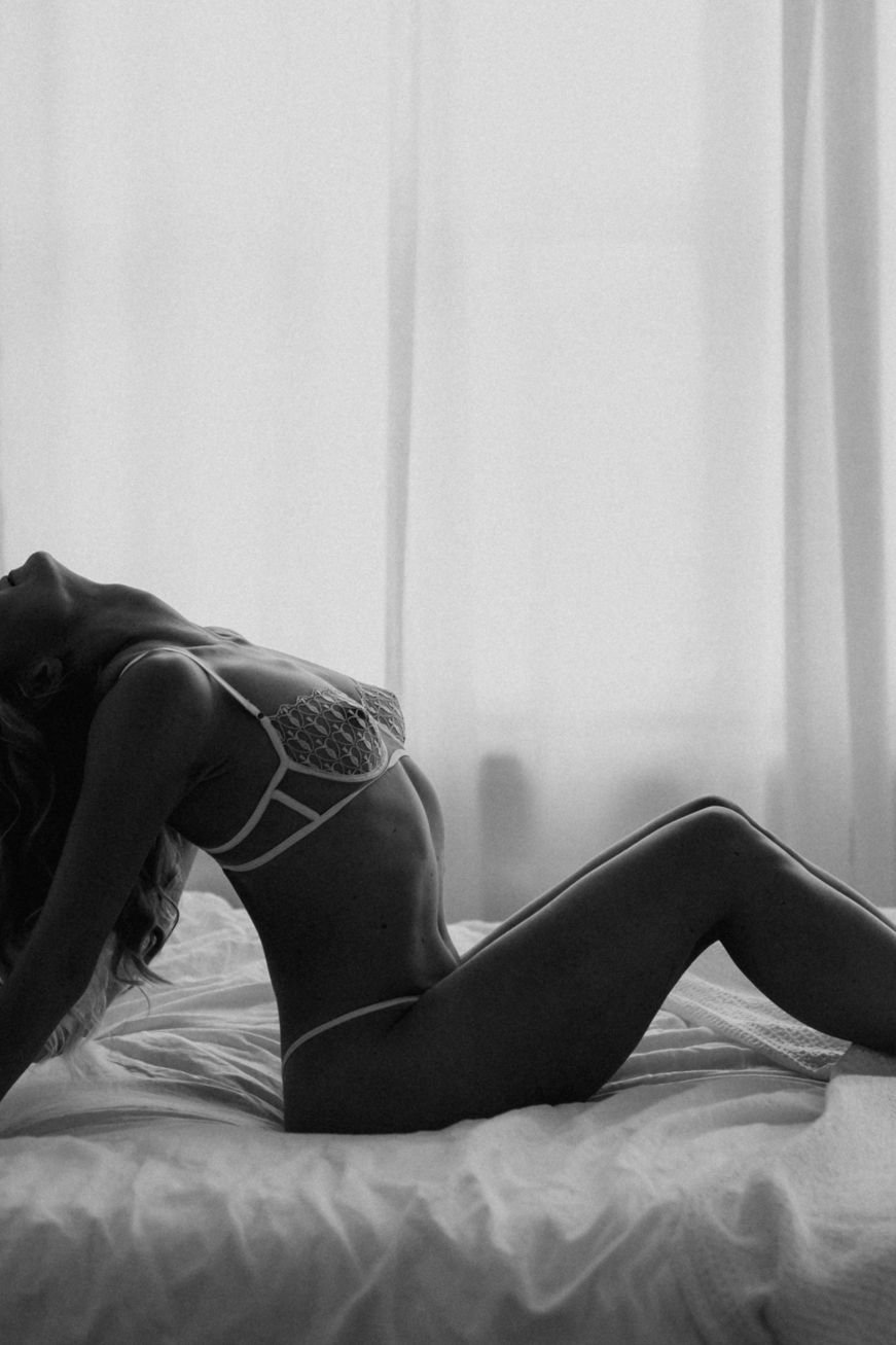 Black and white photo of woman in strappy lingerie posing on white bed.
