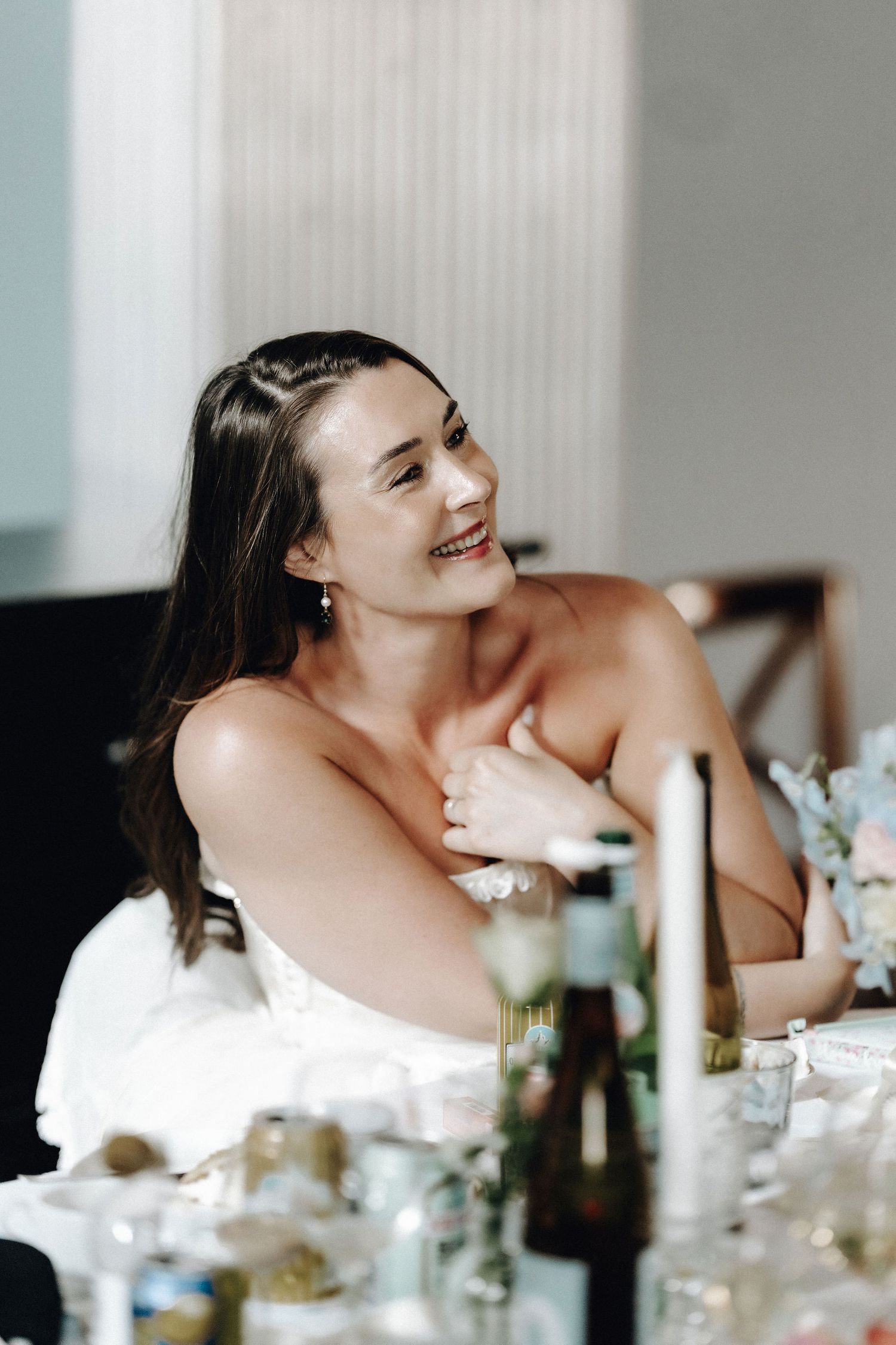 Smiling bride in white strapless gown seated at wedding reception table with floral arrangements.