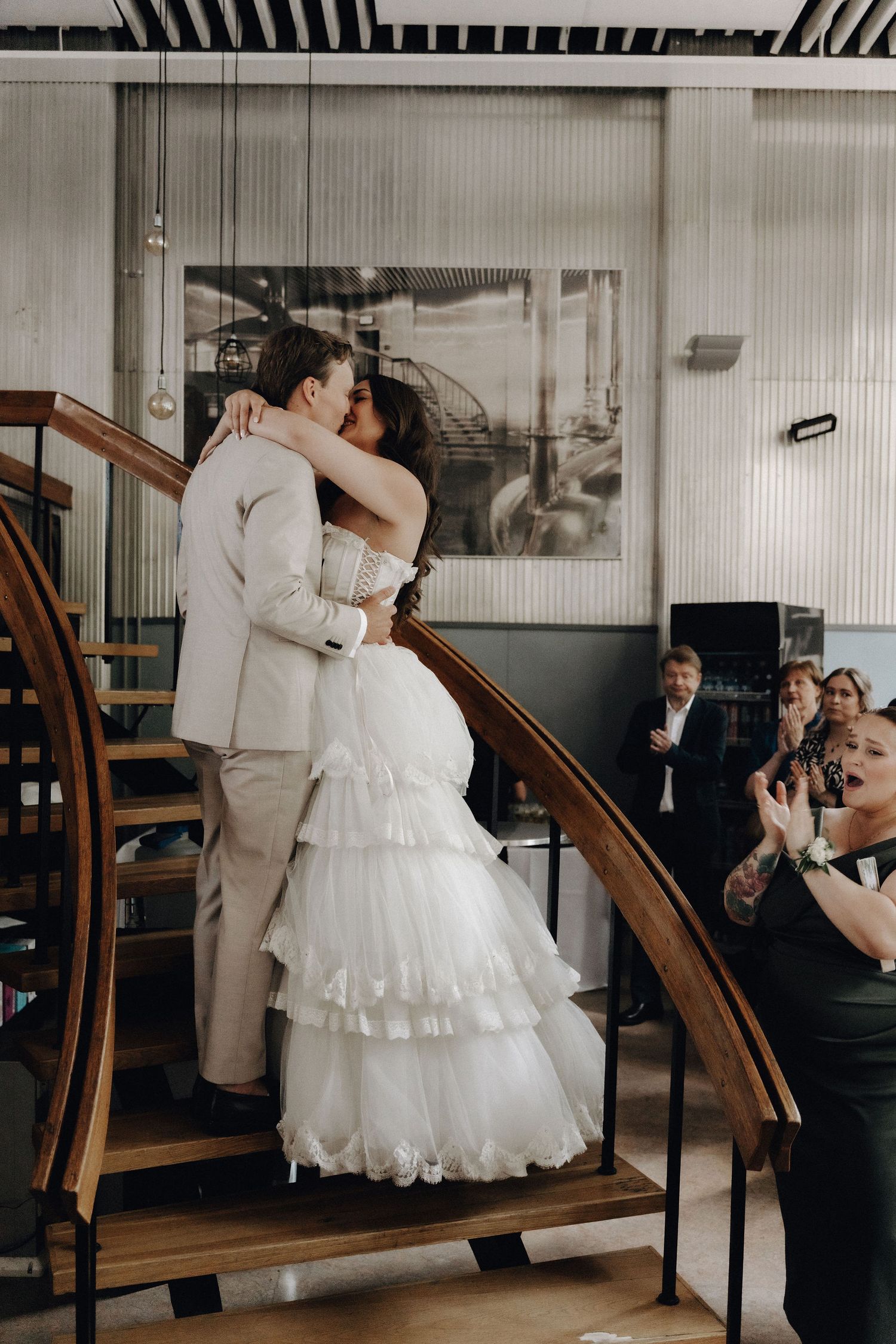 Newlyweds share their first kiss on a curved wooden staircase as guests applaud during wedding reception.