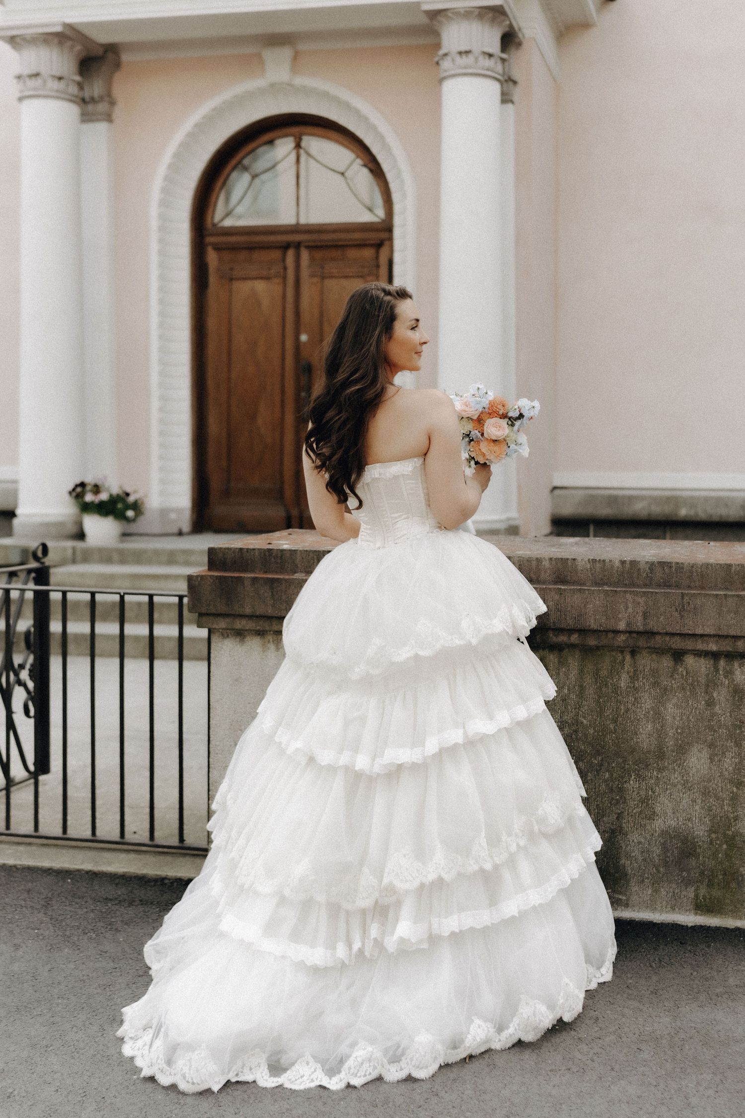 Elegant bride in a strapless white layered ruffle ball gown holding a bouquet, standing before a classic doorway.