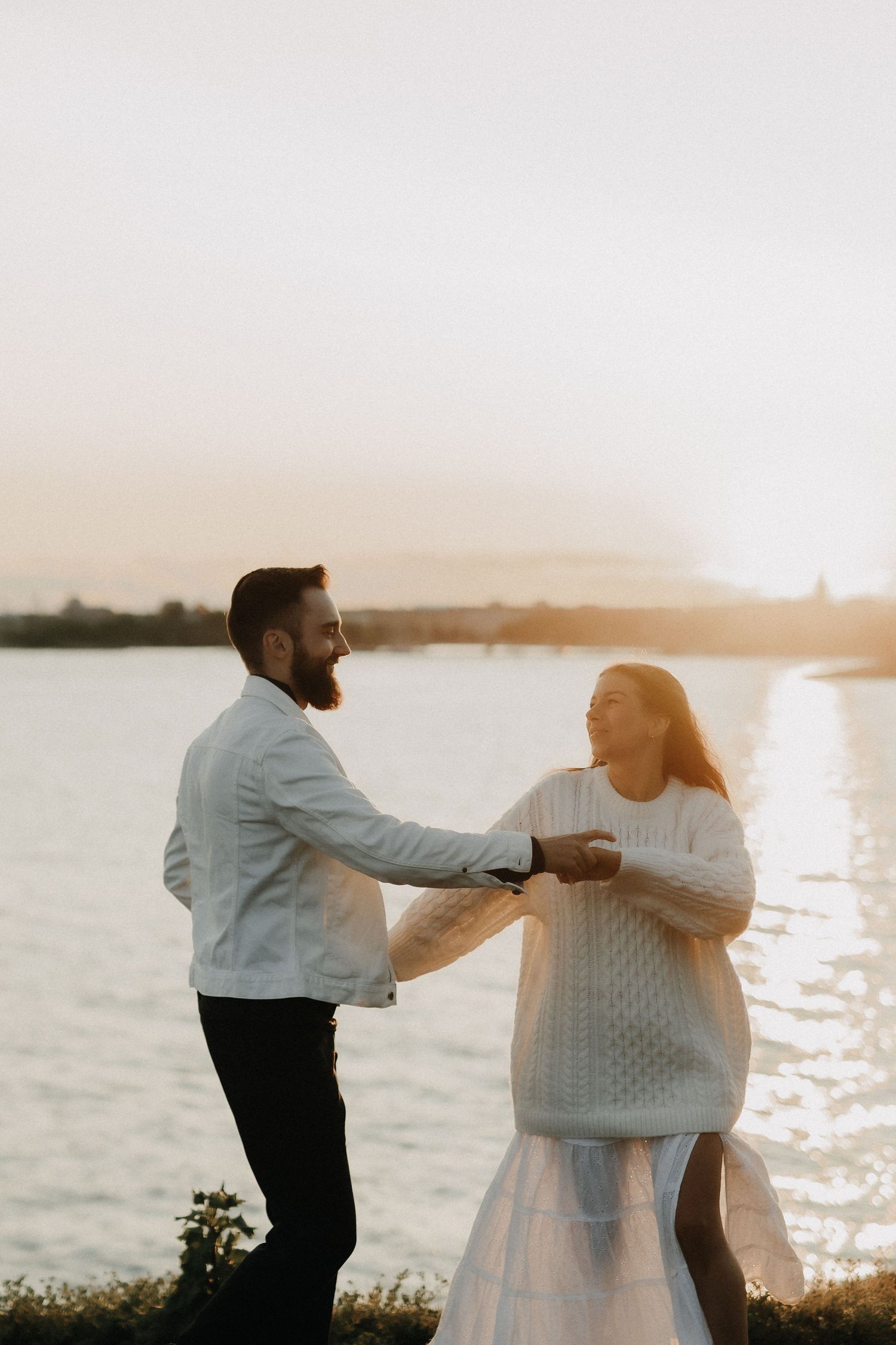 Joyful couple dancing and holding hands by a shimmering lake during a warm golden sunset.