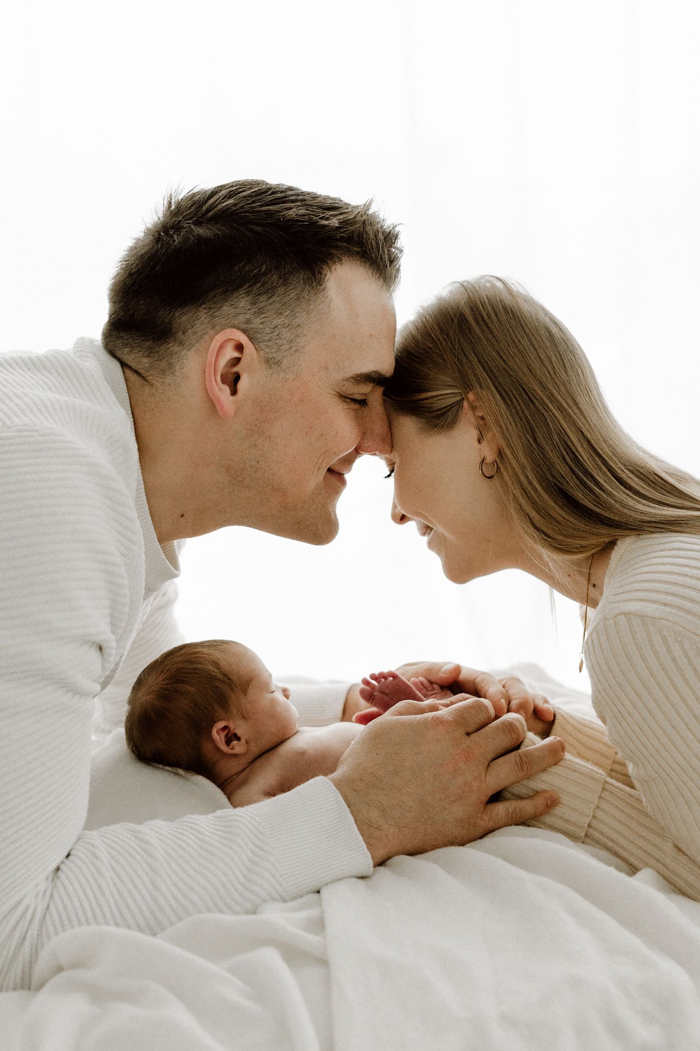 Loving parents touch foreheads while holding their sleeping newborn baby together on a white blanket.