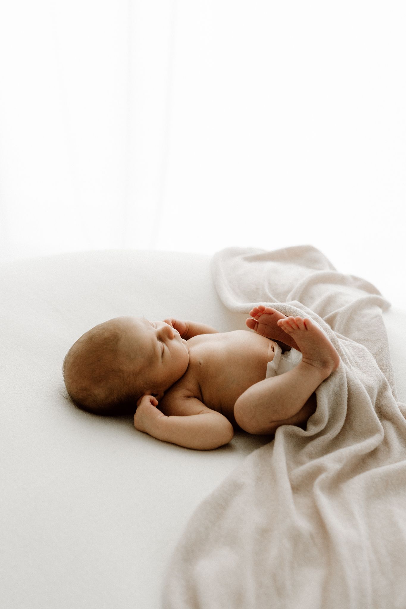 Serene newborn baby sleeping on white surface with light beige blanket in natural light.
