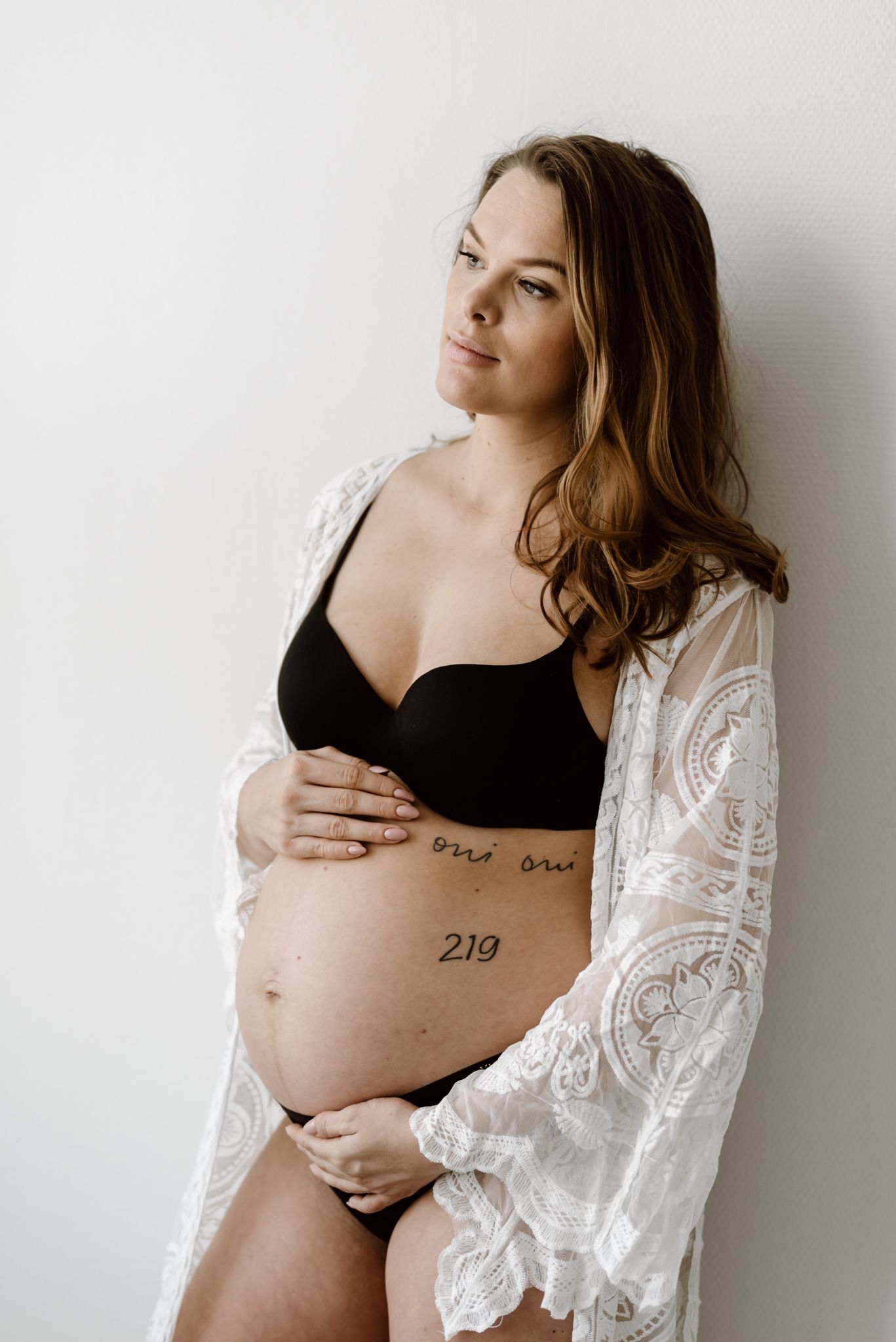 Pregnant woman in black lingerie and white lace robe cradling tattooed baby bump indoors.