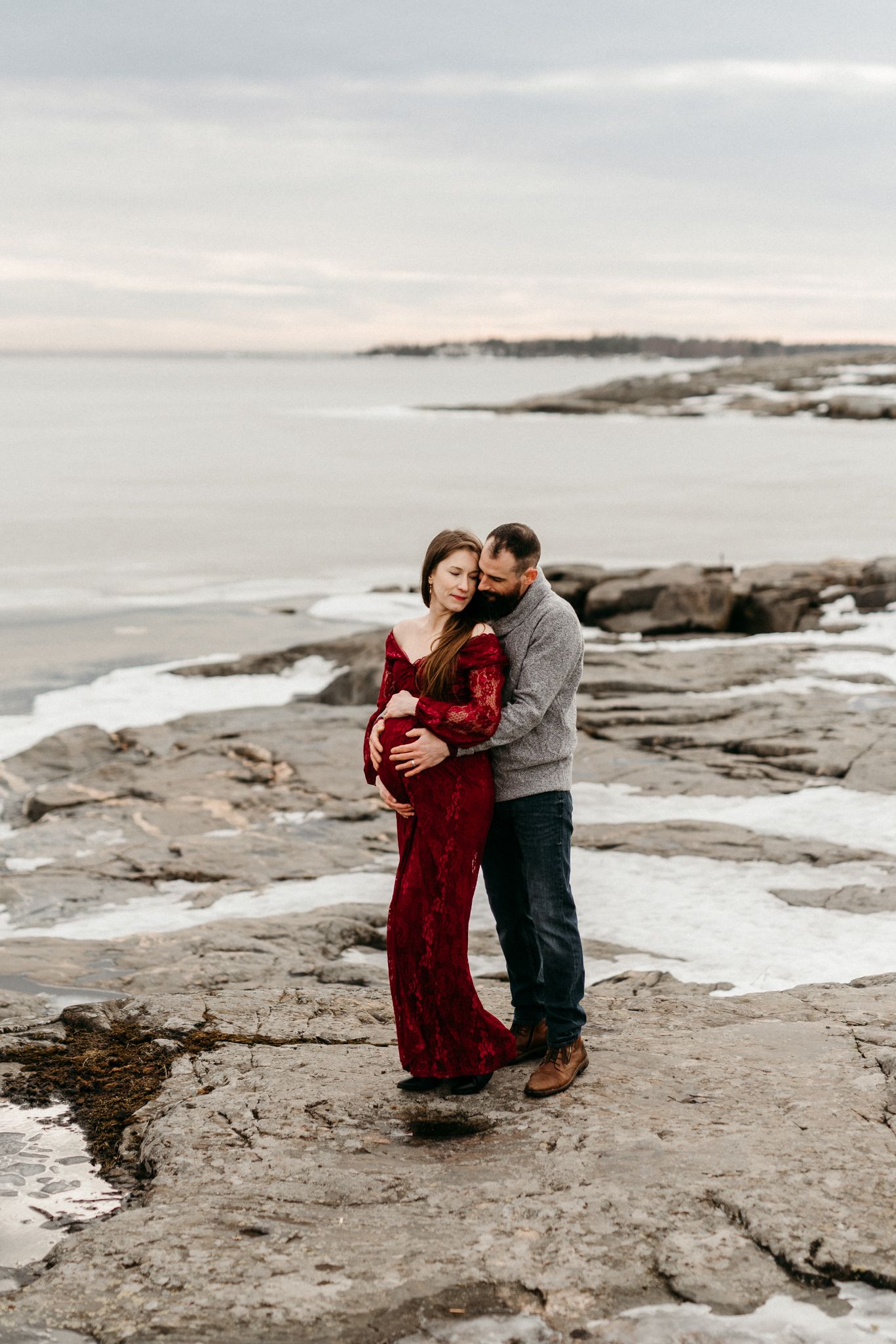 Pregnant couple embracing on rocky winter coastline, woman in red lace gown, man in gray sweater at dusk.