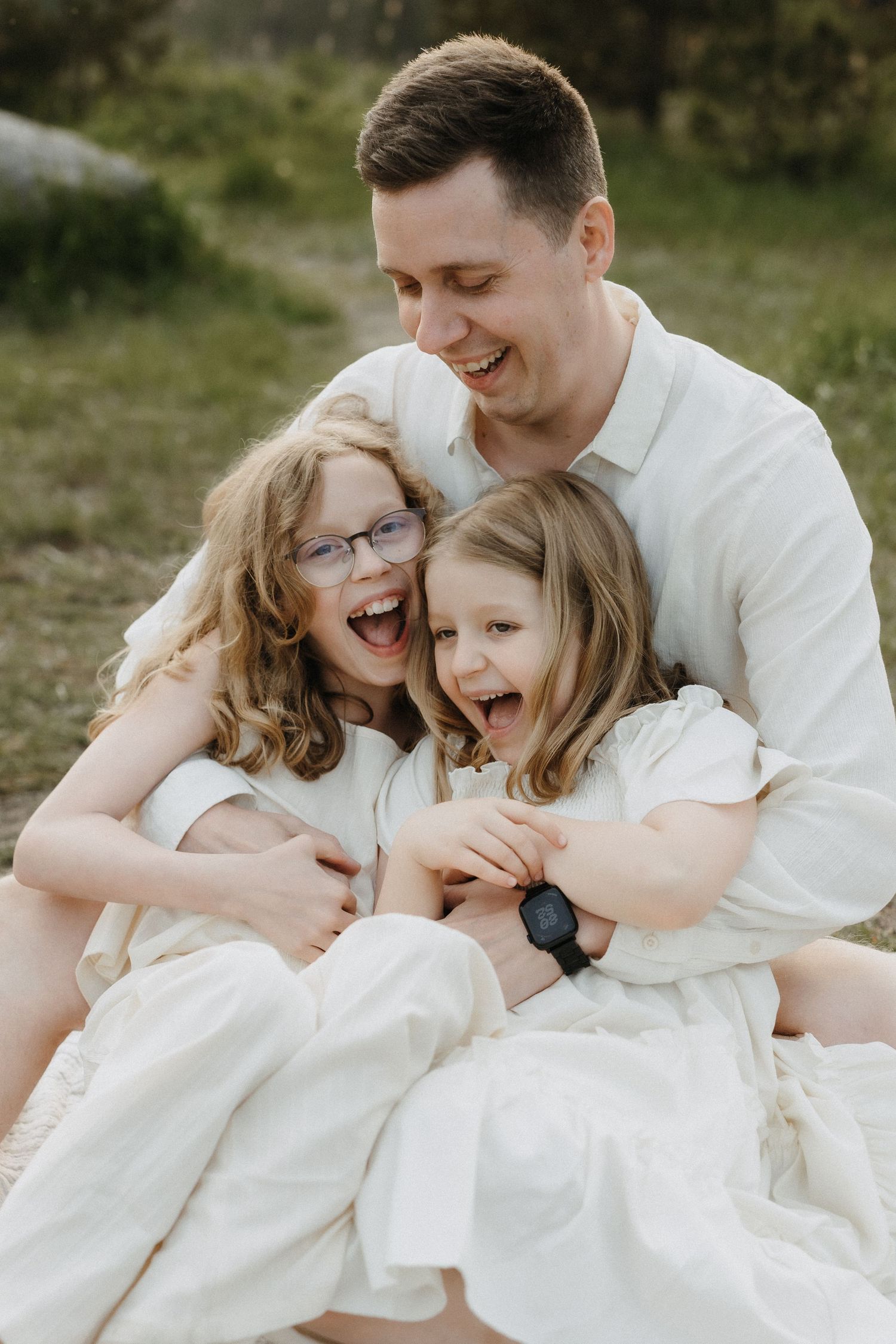 A joyful father sits outdoors hugging his two laughing daughters in matching white dresses.