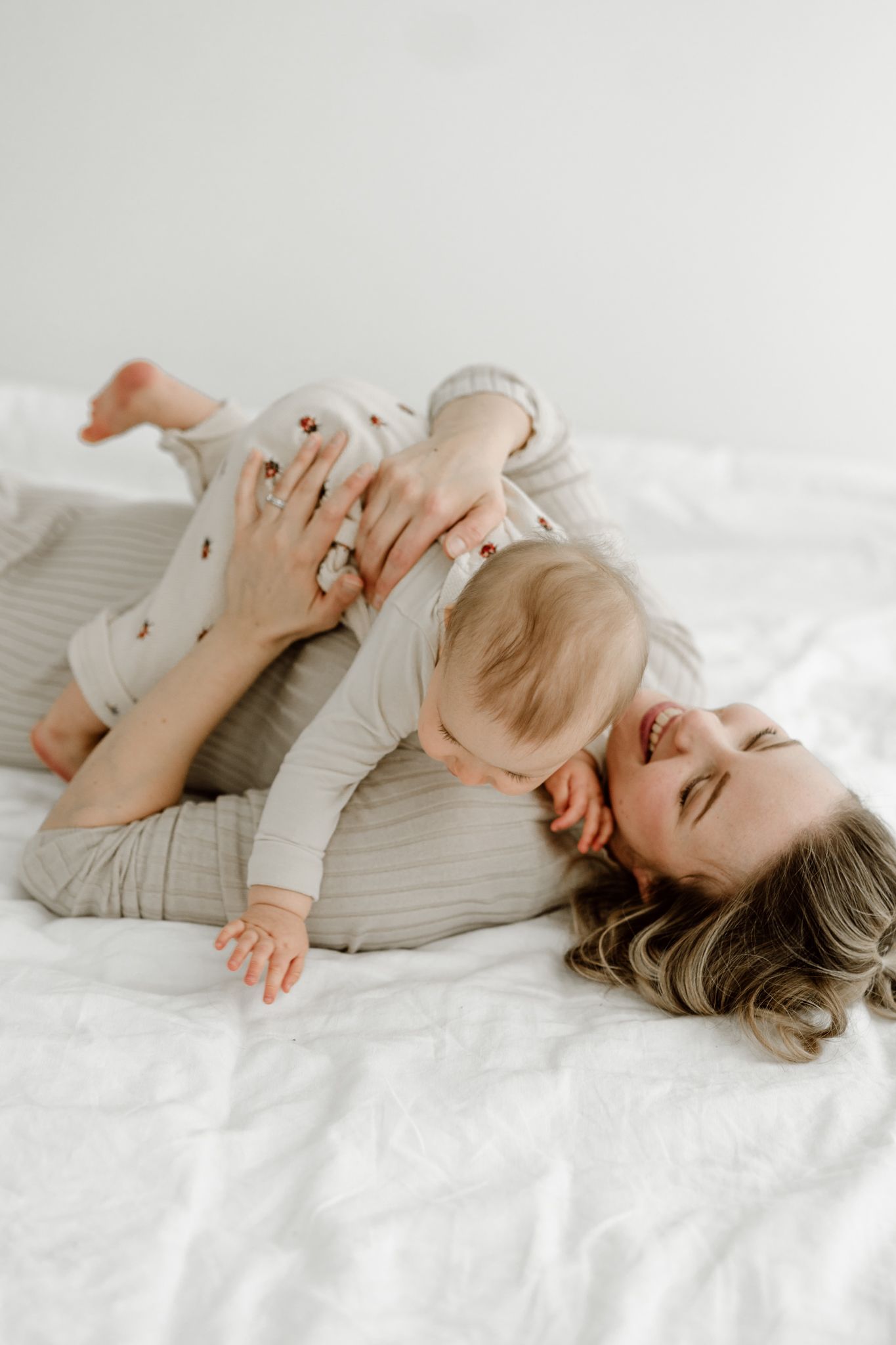 Playful mother lying on a white bed cuddling and laughing with her happy baby.