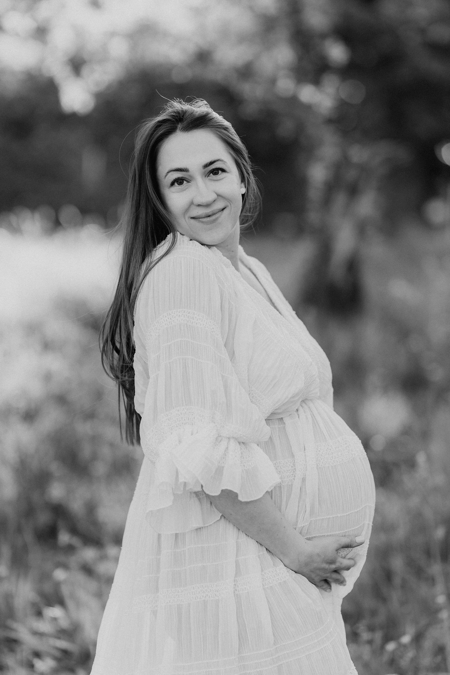 Smiling pregnant woman in white dress holding baby bump in black and white outdoor portrait.