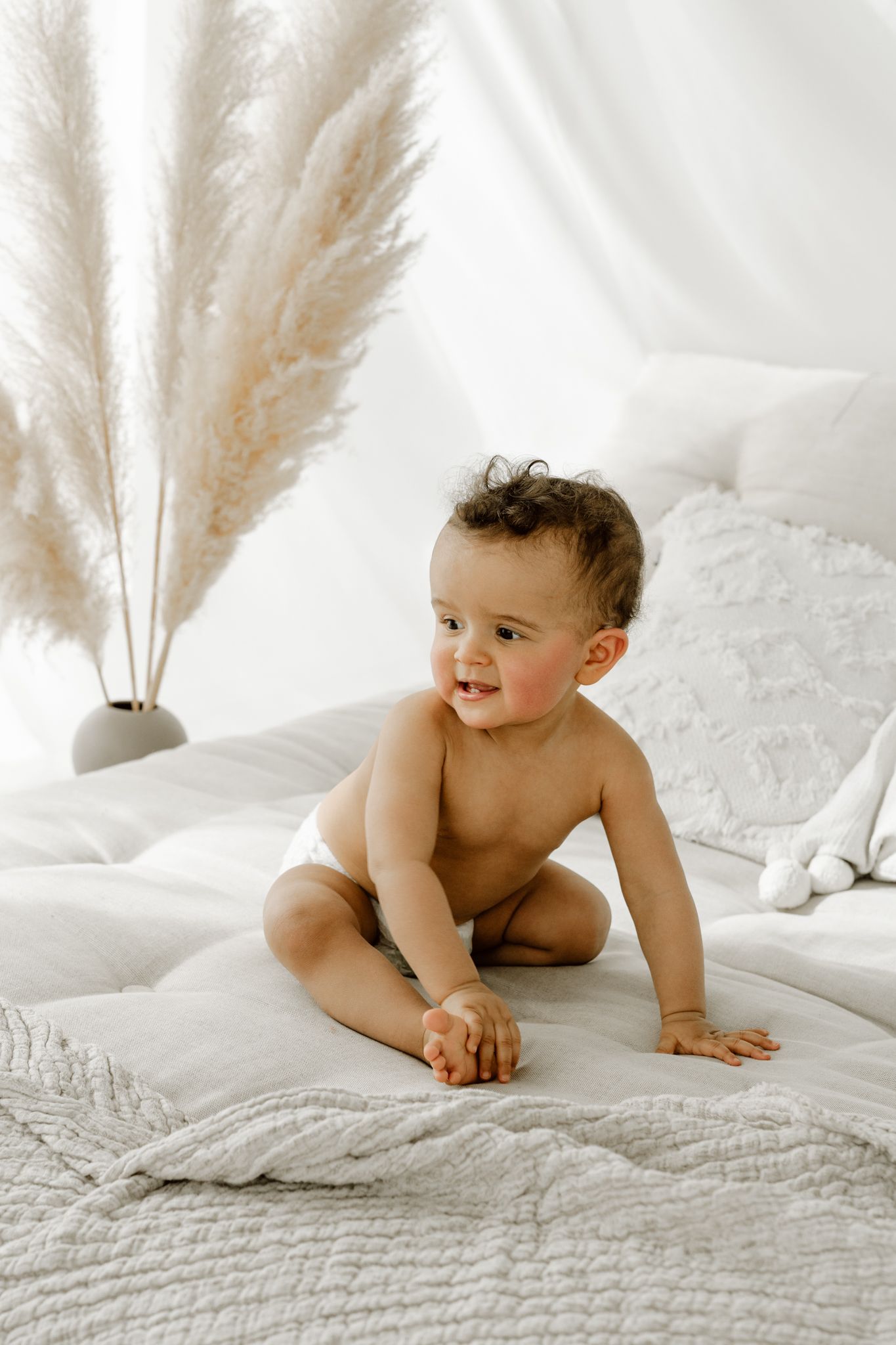 Cheerful baby in diaper sitting on white quilted bed near pampas grass decoration.