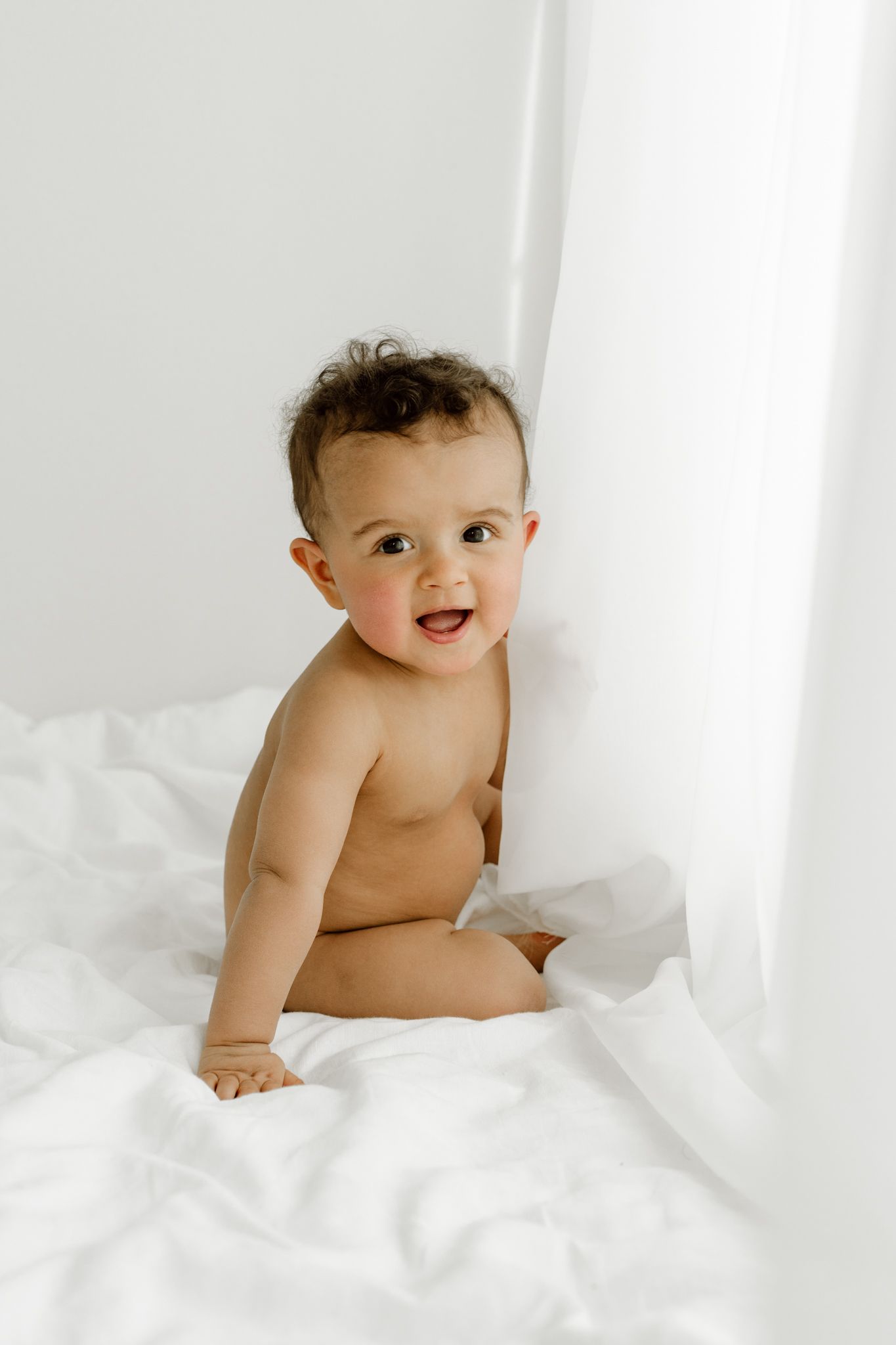 Happy shirtless baby sitting on white sheets smiling at camera near white curtains.