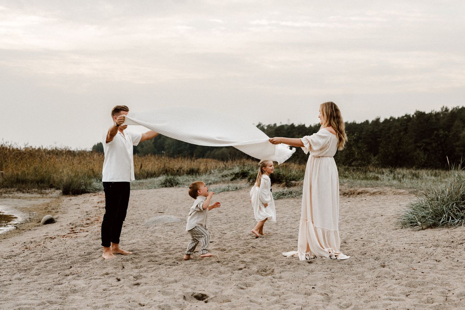 A family of four plays with a white blanket on a sandy beach at sunset.
