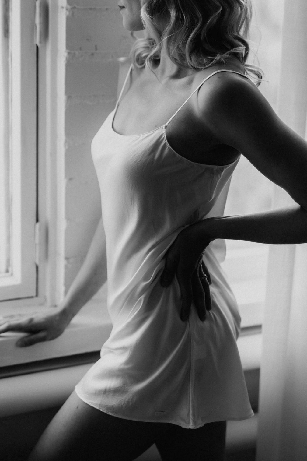 Black and white photo of woman in white slip dress posing by a window.