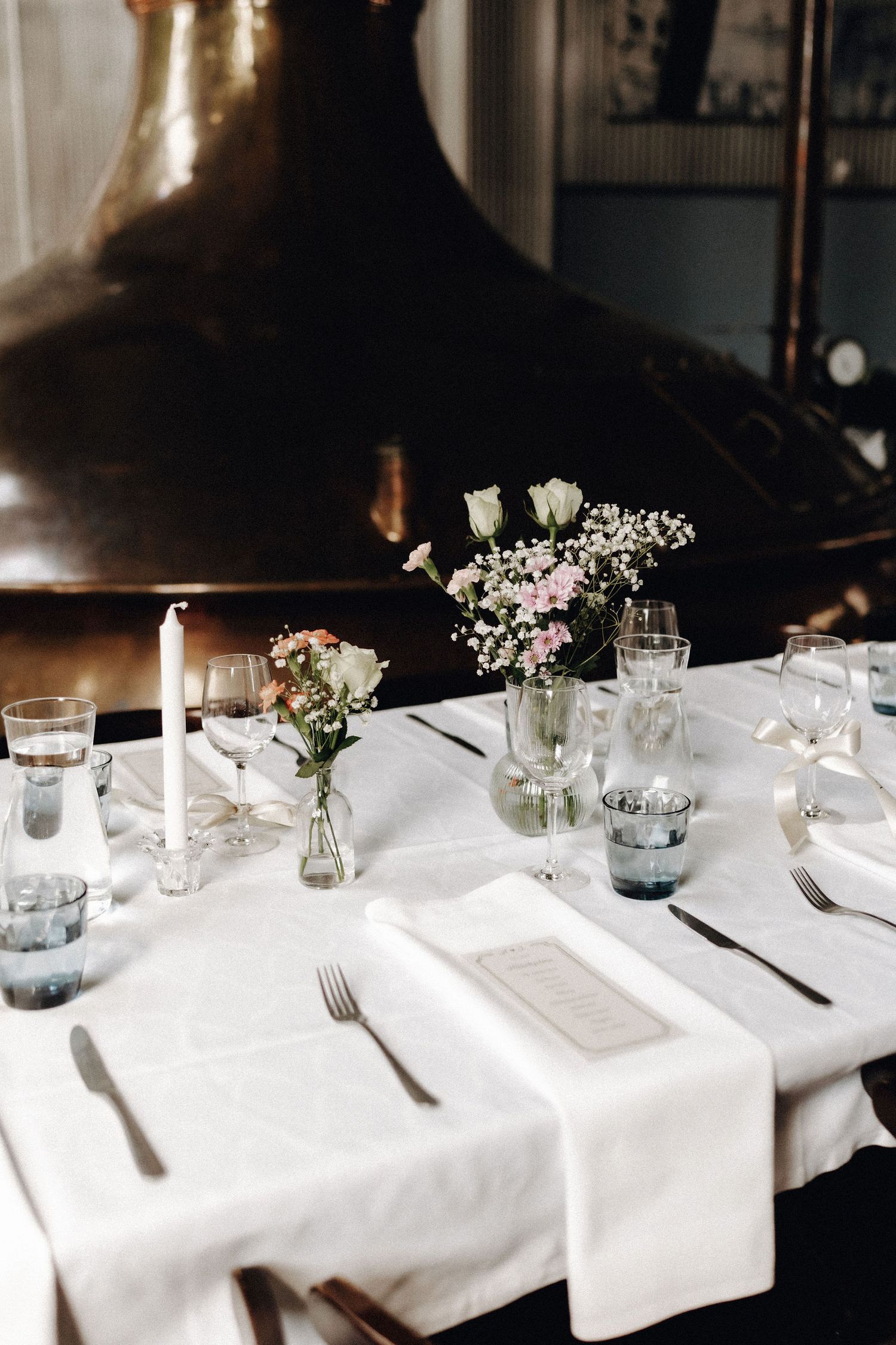 Elegant wedding reception table with white linens, candles, wildflowers, and glassware in brewery venue.