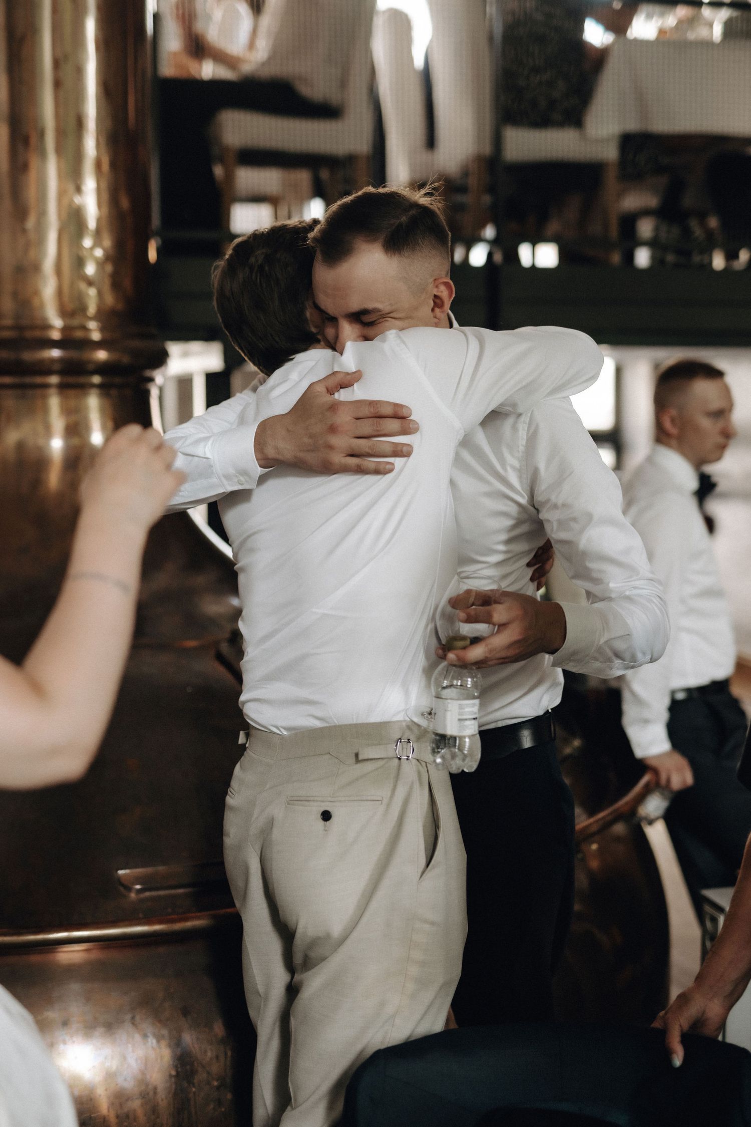 Two men in white shirts sharing warm embrace near copper brewery tanks at wedding reception.