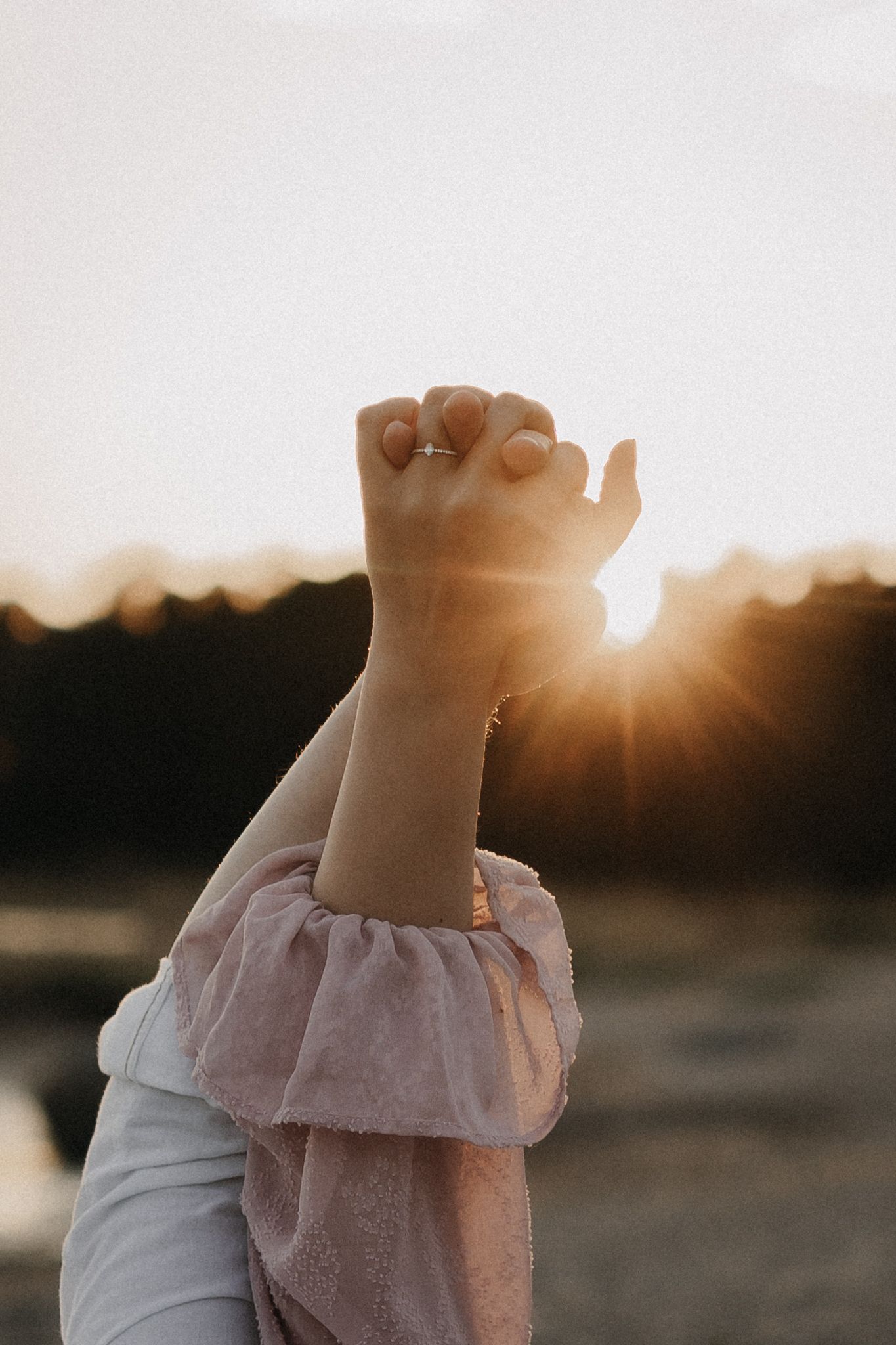 Hands raised against glowing sunset sky, woman wearing pink dress sleeve and engagement ring.