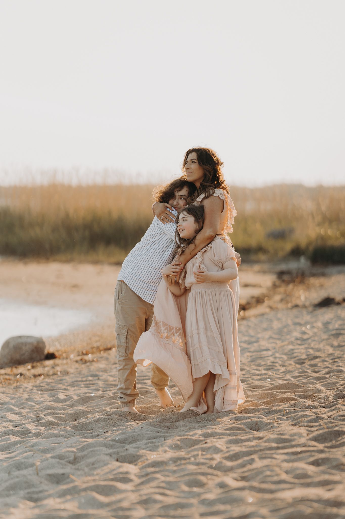 A family of three shares a warm embrace on a sandy beach at golden hour, wearing soft neutral tones.
