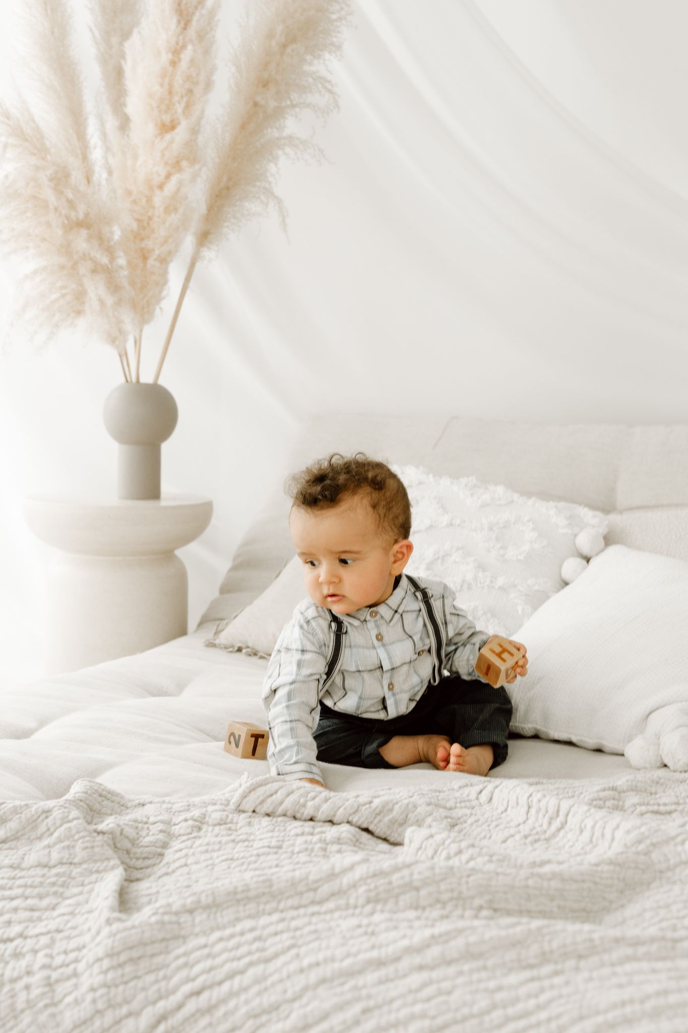 Curious baby boy in plaid shirt sitting on white quilted bed with wooden toy blocks.
