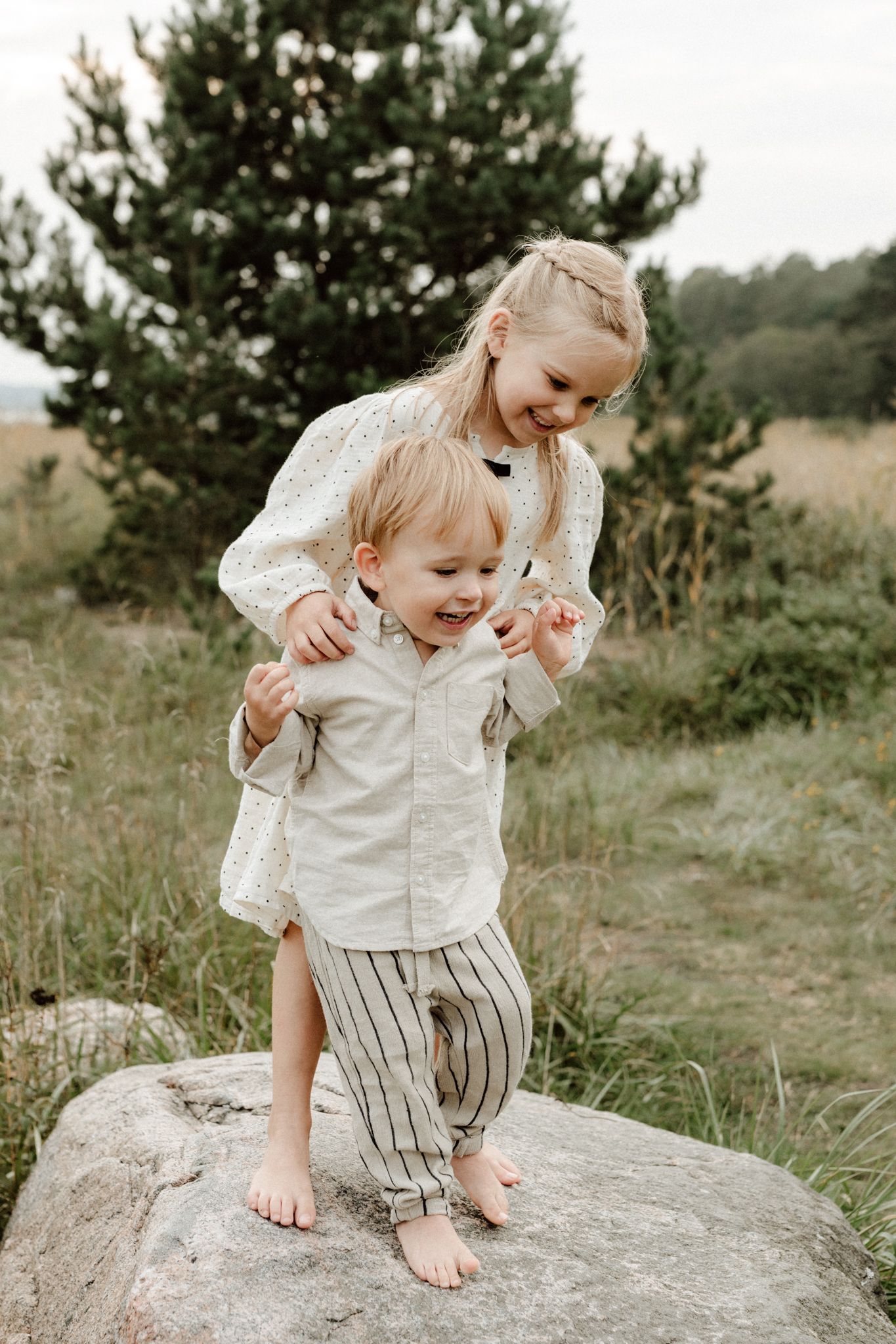Smiling girl helping laughing toddler brother stand on large rock in grassy field.