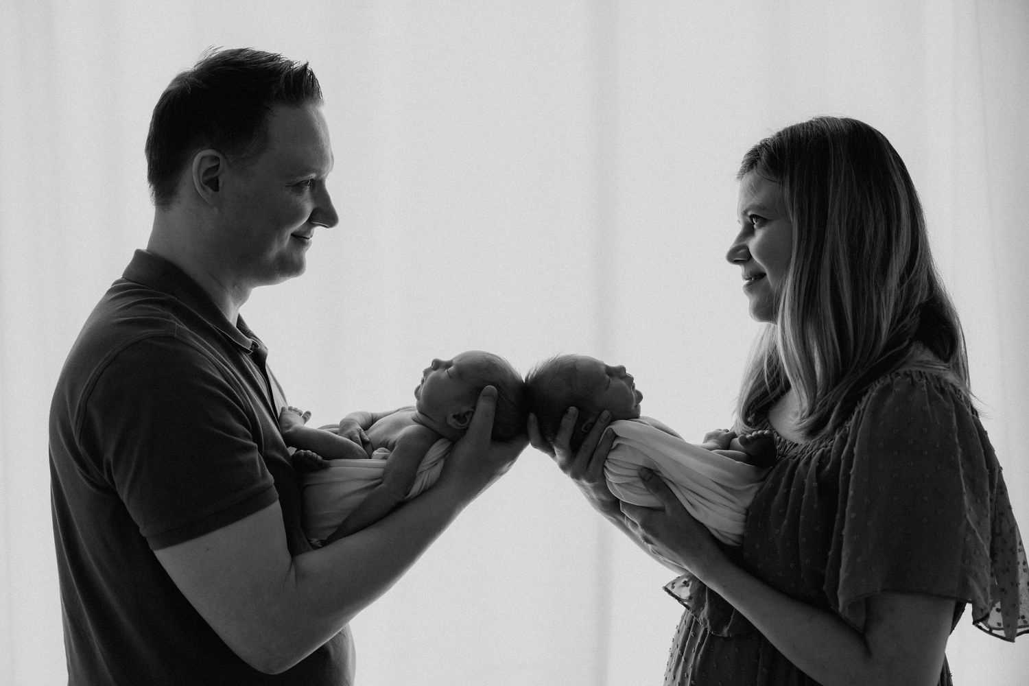 Black and white photo of couple facing each other, each holding a newborn twin baby.