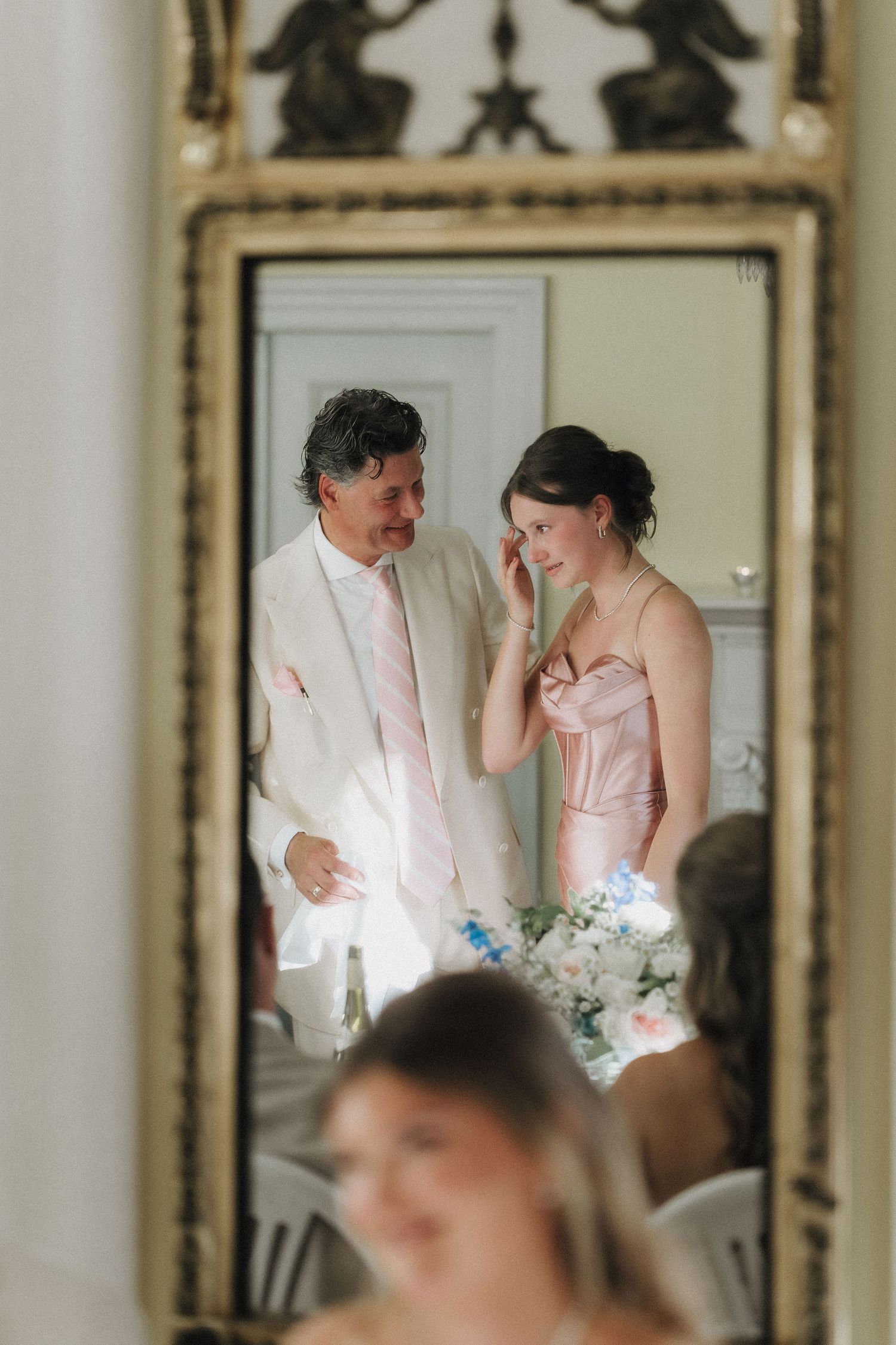 Reflection in ornate gold mirror of man comforting emotional woman in pink dress at wedding.