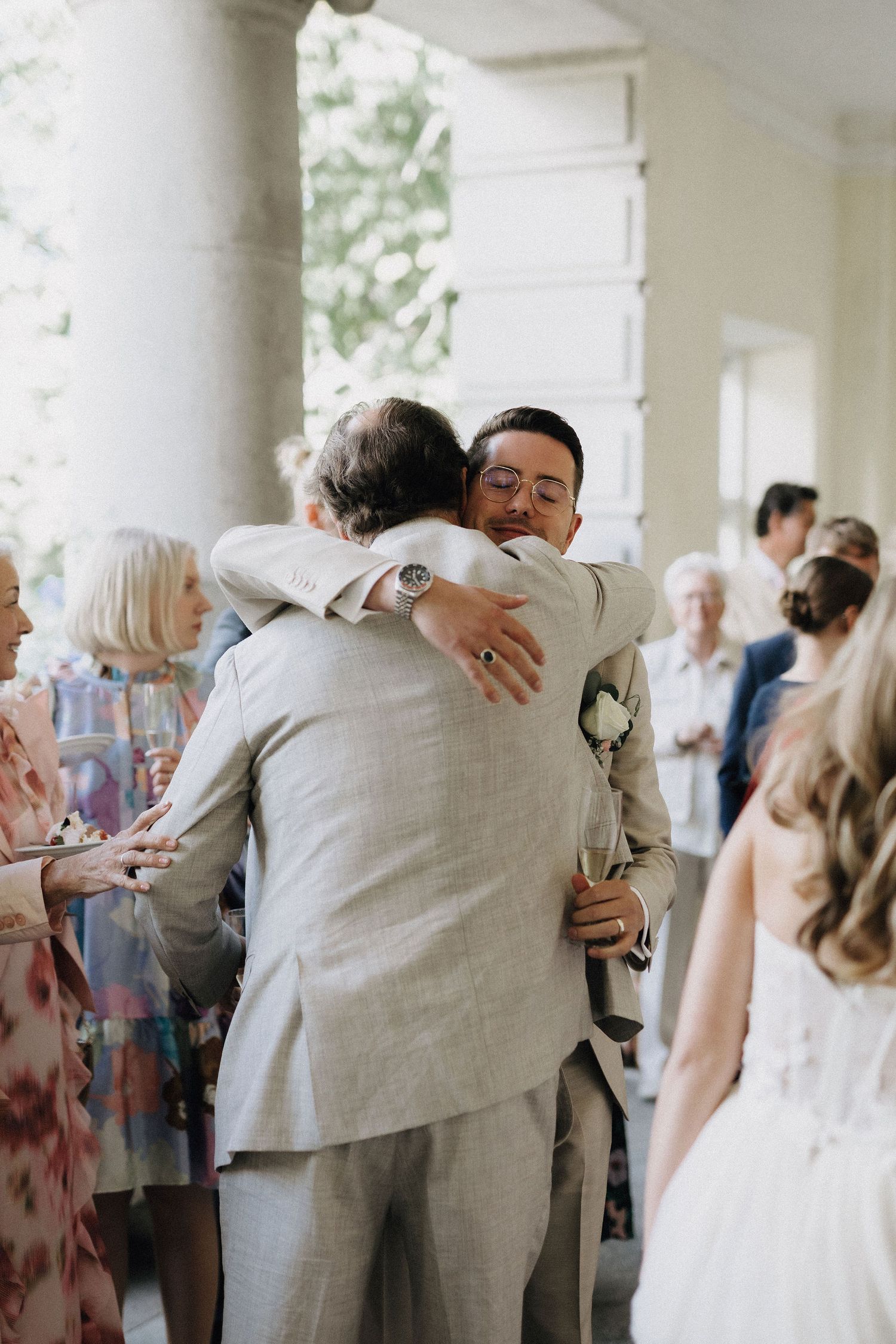 Groom in beige suit warmly embraces a wedding guest outdoors at elegant venue reception.