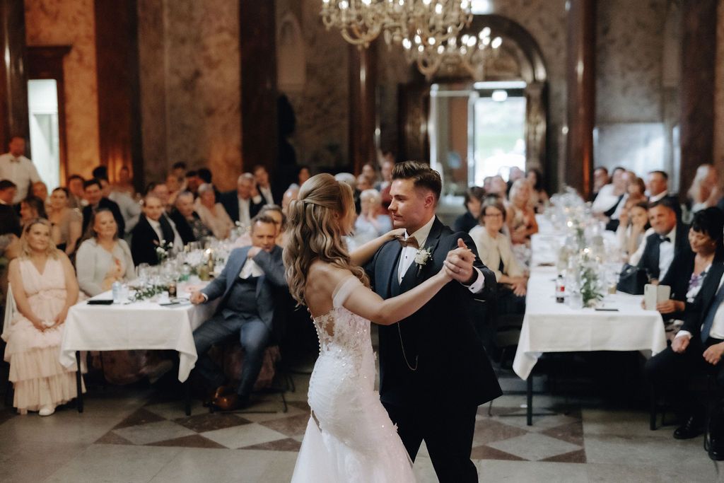 Bride and groom smiling during first dance in ornate ballroom with guests watching nearby.