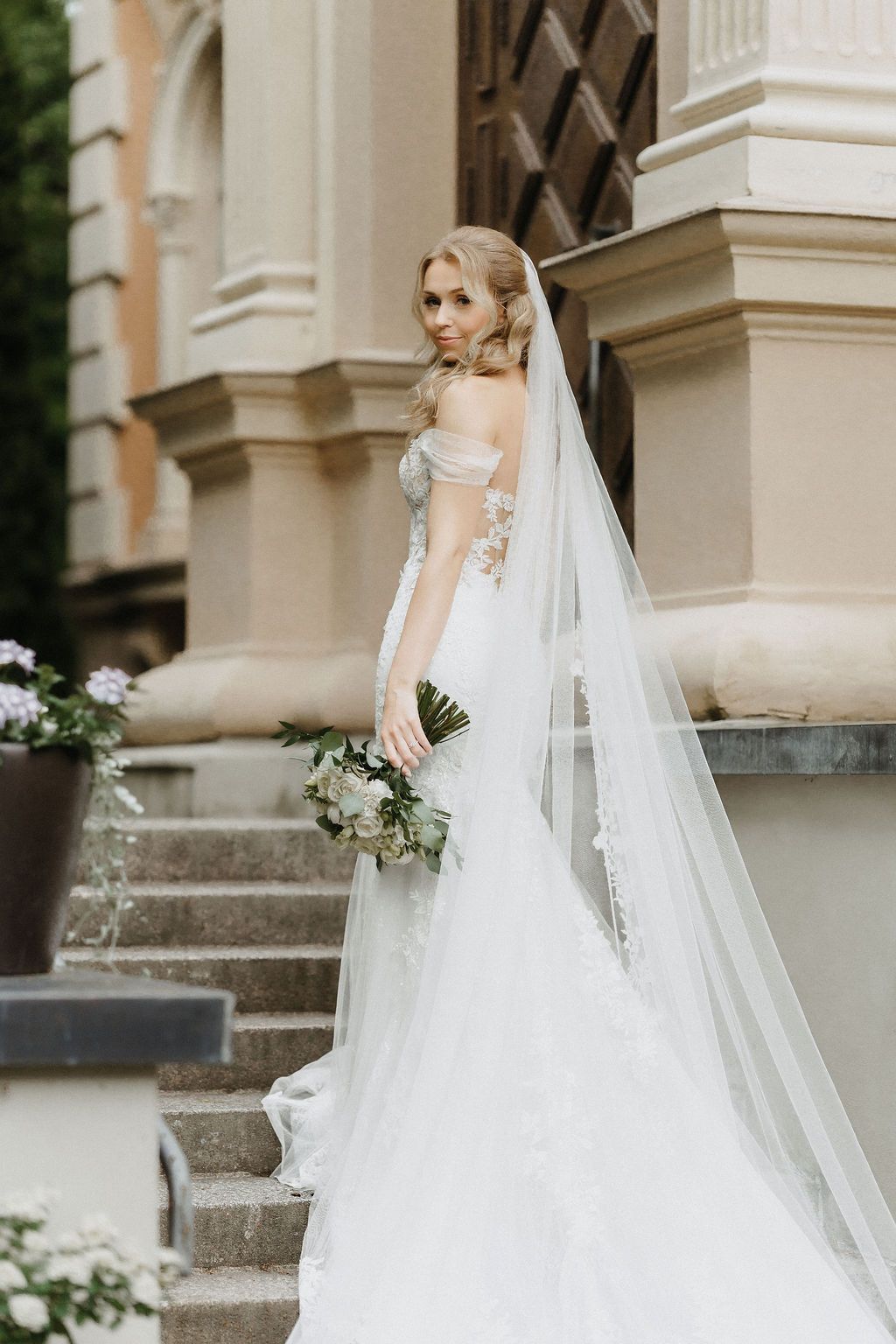 Elegant bride in off-shoulder lace gown with cathedral veil holds bouquet on stone steps of classic building.