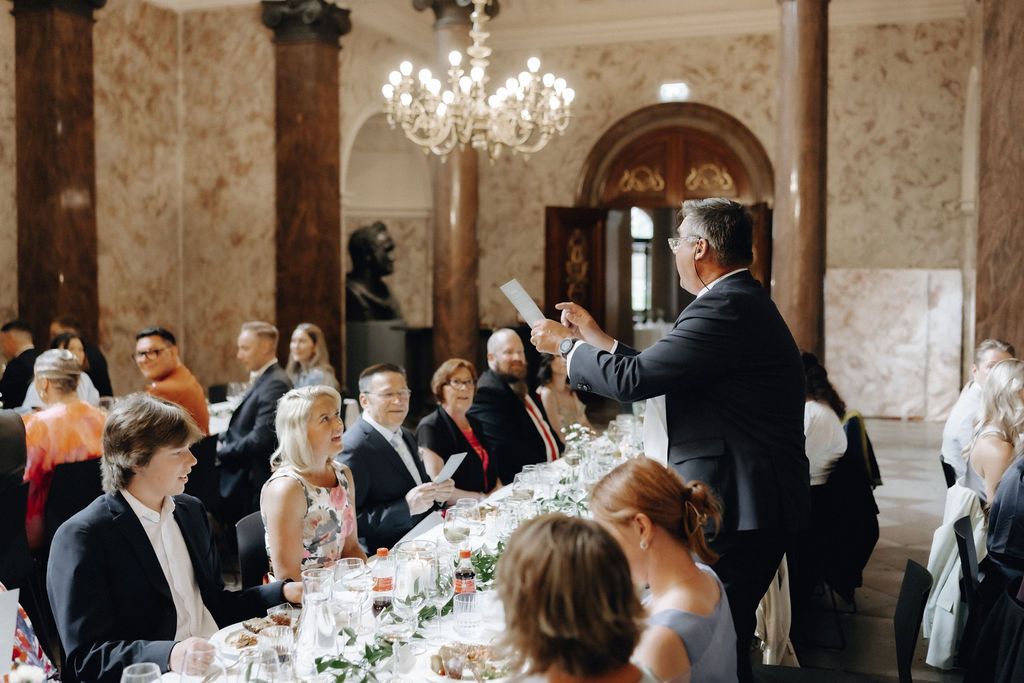 Man reading speech at wedding banquet in luxurious marble columned hall with crystal chandelier.