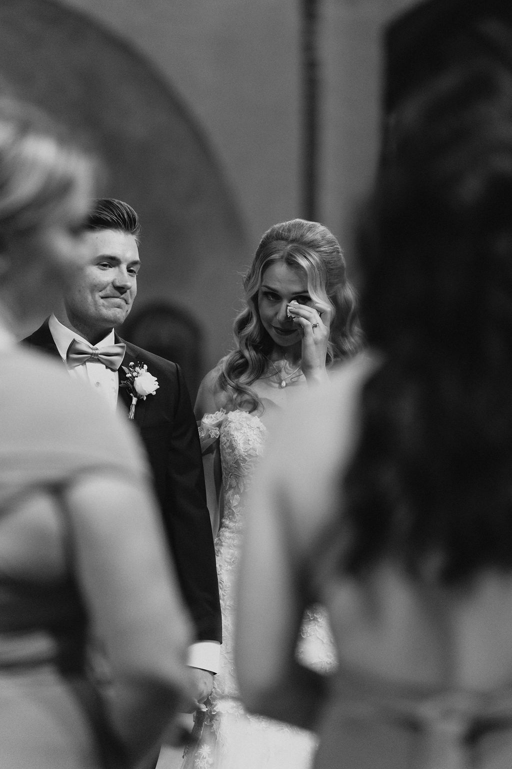 Emotional bride wiping tears during wedding ceremony while groom smiles in black tuxedo with bow tie.