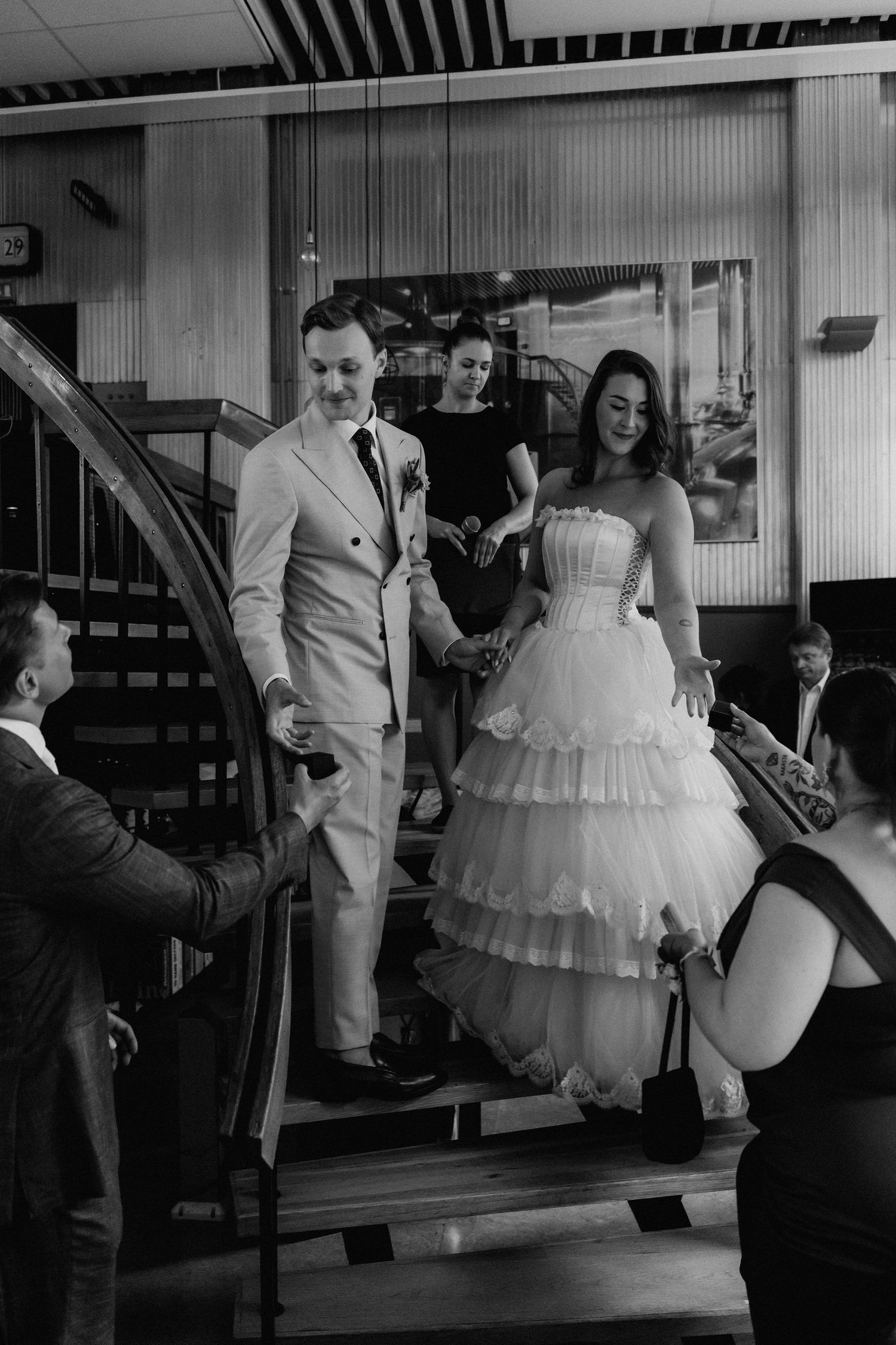 Newlyweds descend a curved wooden staircase hand in hand surrounded by celebrating wedding guests.