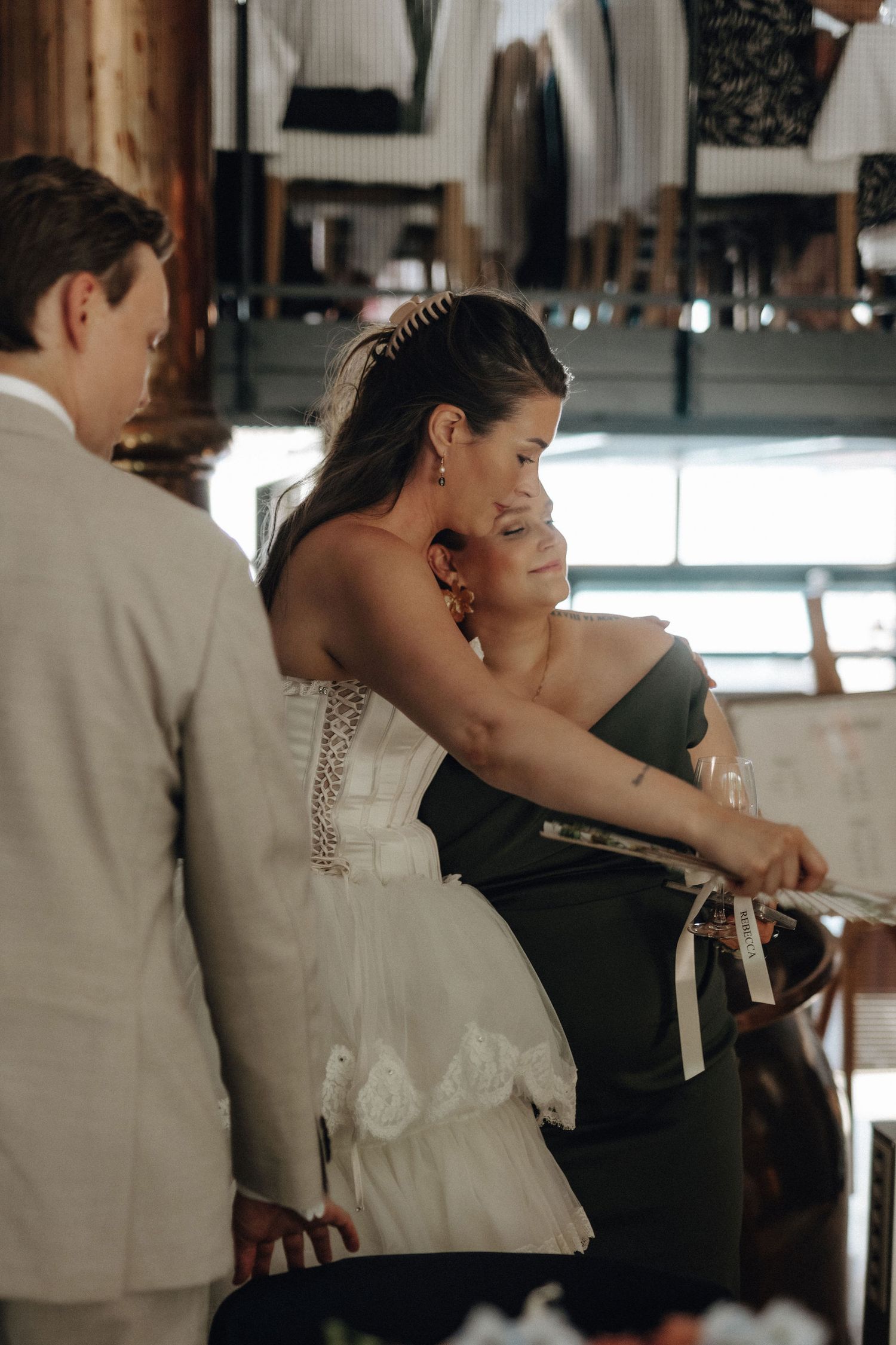 Bride in white ruffled gown embracing bridesmaid in green dress at wedding reception venue.