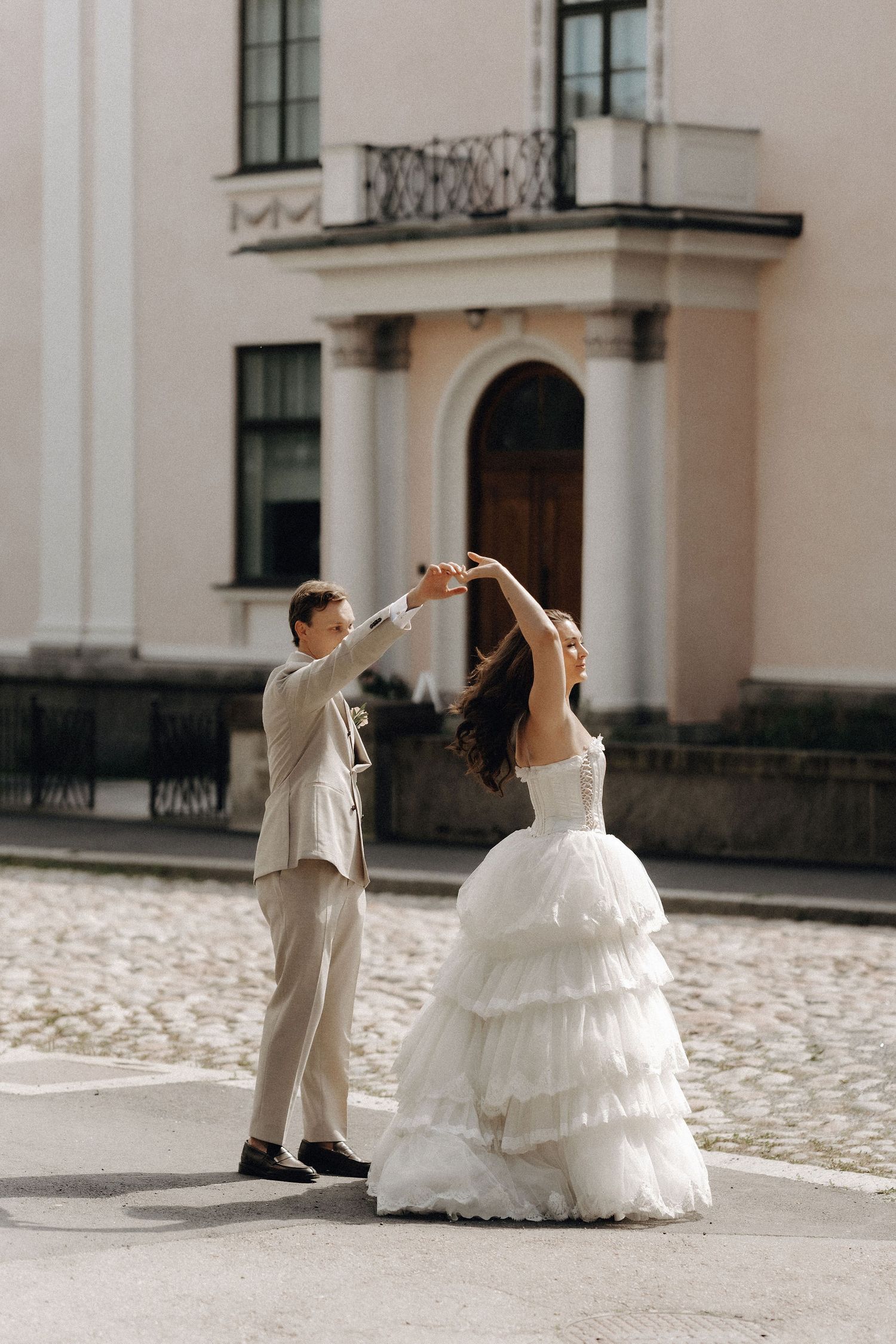 Bride and groom dancing joyfully on a sunny cobblestone plaza outside a classic building.