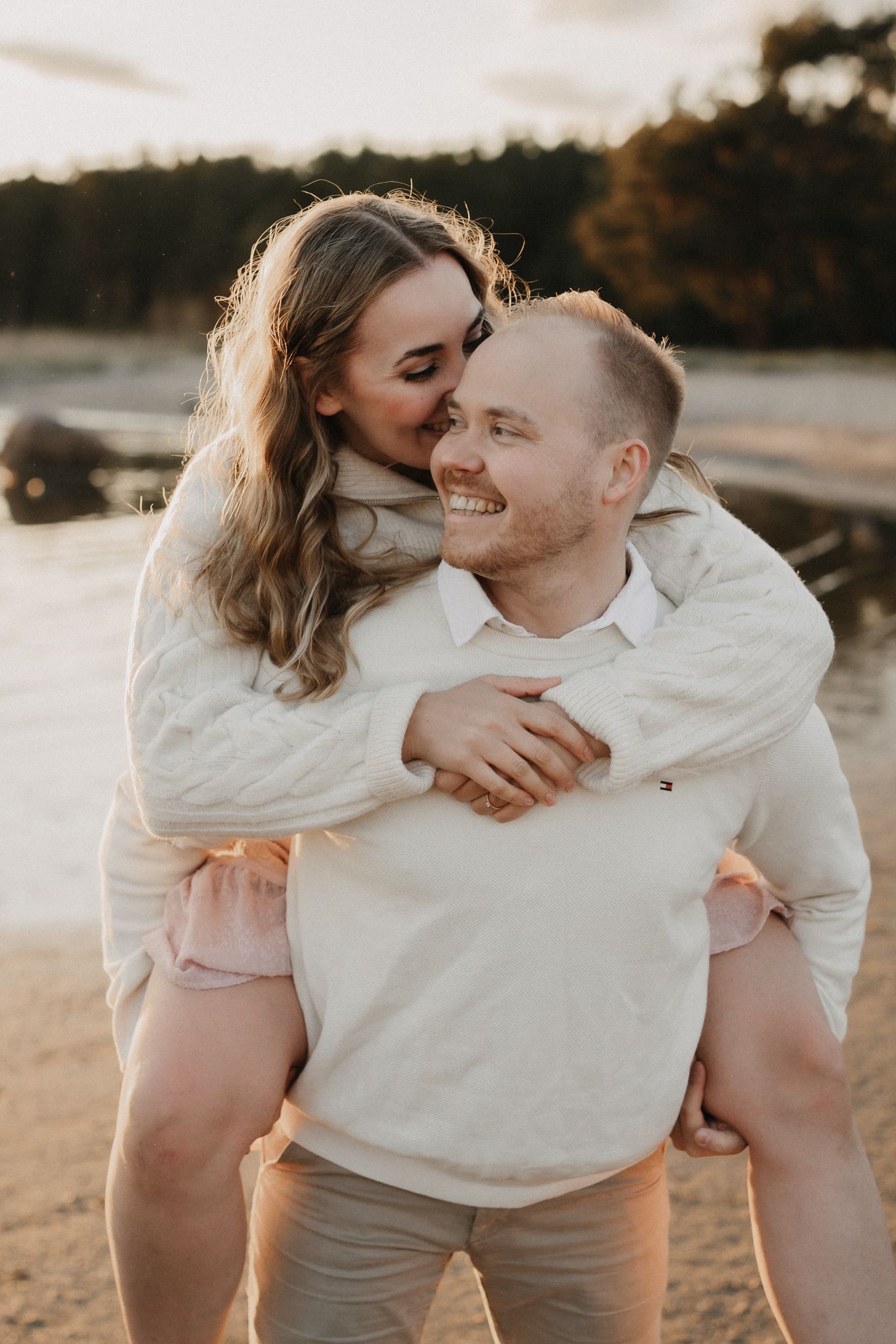 Smiling man gives woman a piggyback ride on a beach at sunset, both laughing joyfully together.