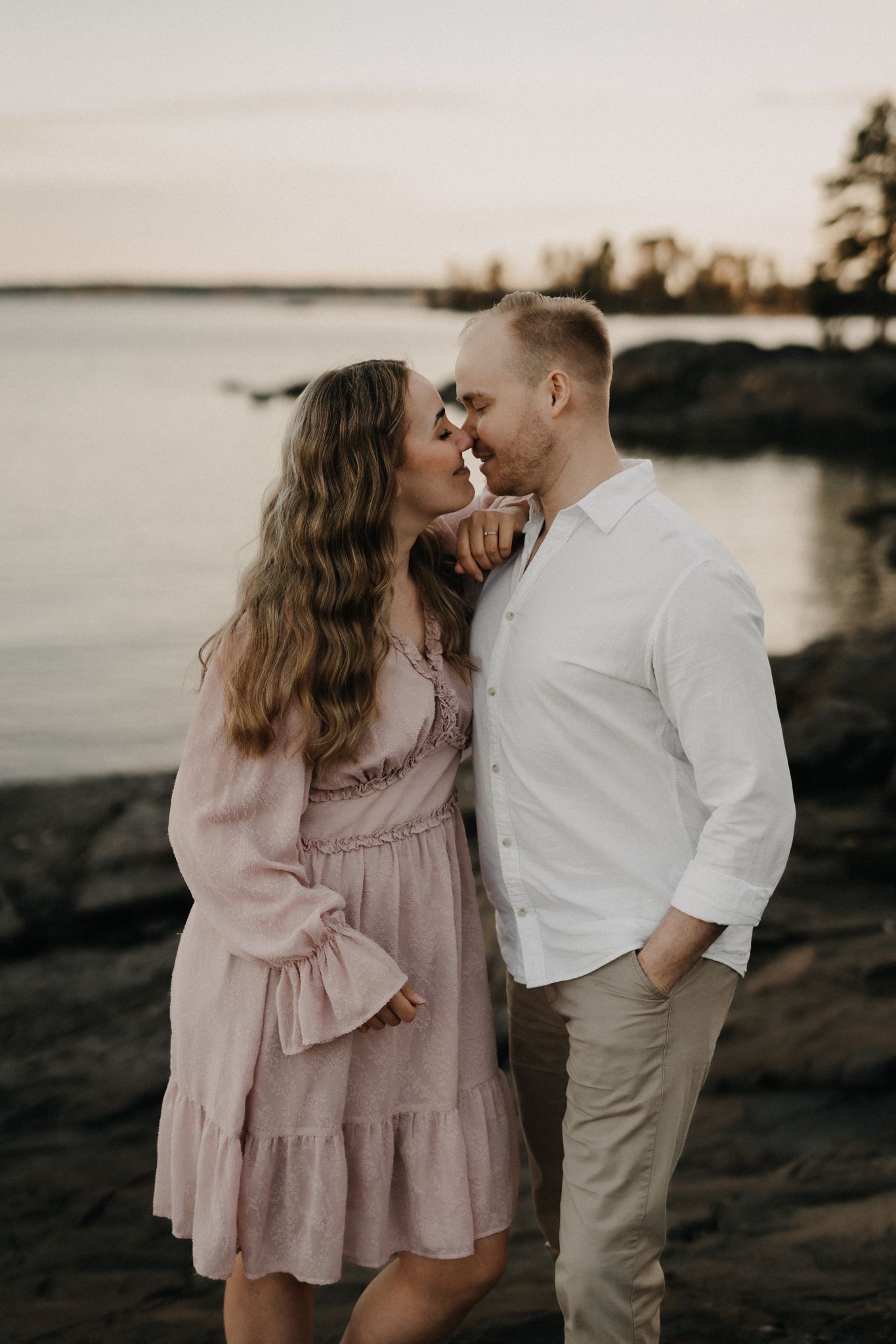 Couple about to kiss near rocky lakeside shore at sunset, woman in pink dress, man in white shirt.