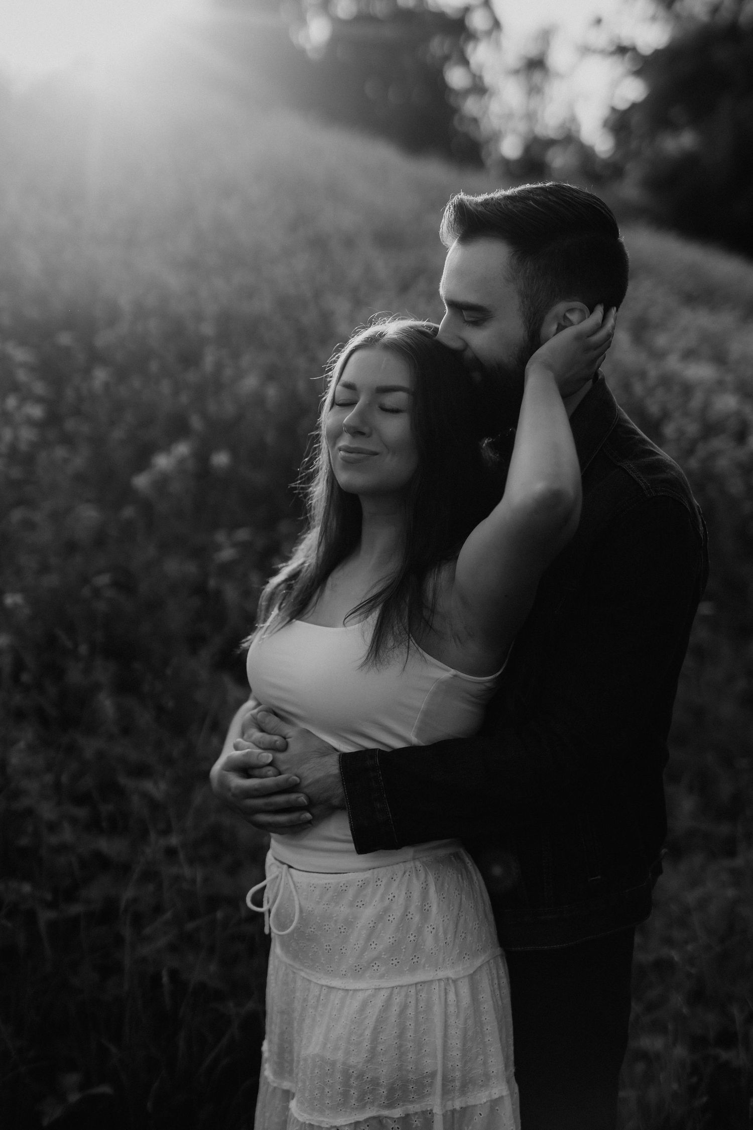 Black and white photo of a couple embracing in a field at sunset, man kissing womans head.