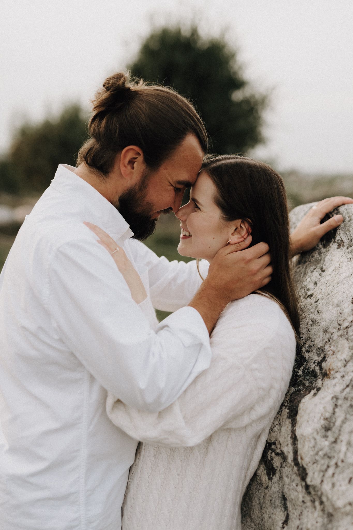 Couple in white outfits sharing a tender moment leaning against a large rock outdoors.