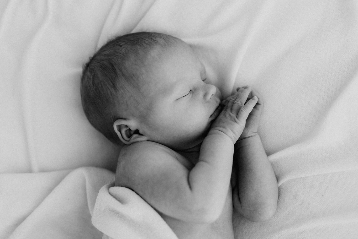 Close-up black and white photo of a sleeping newborn baby with hands folded near face.