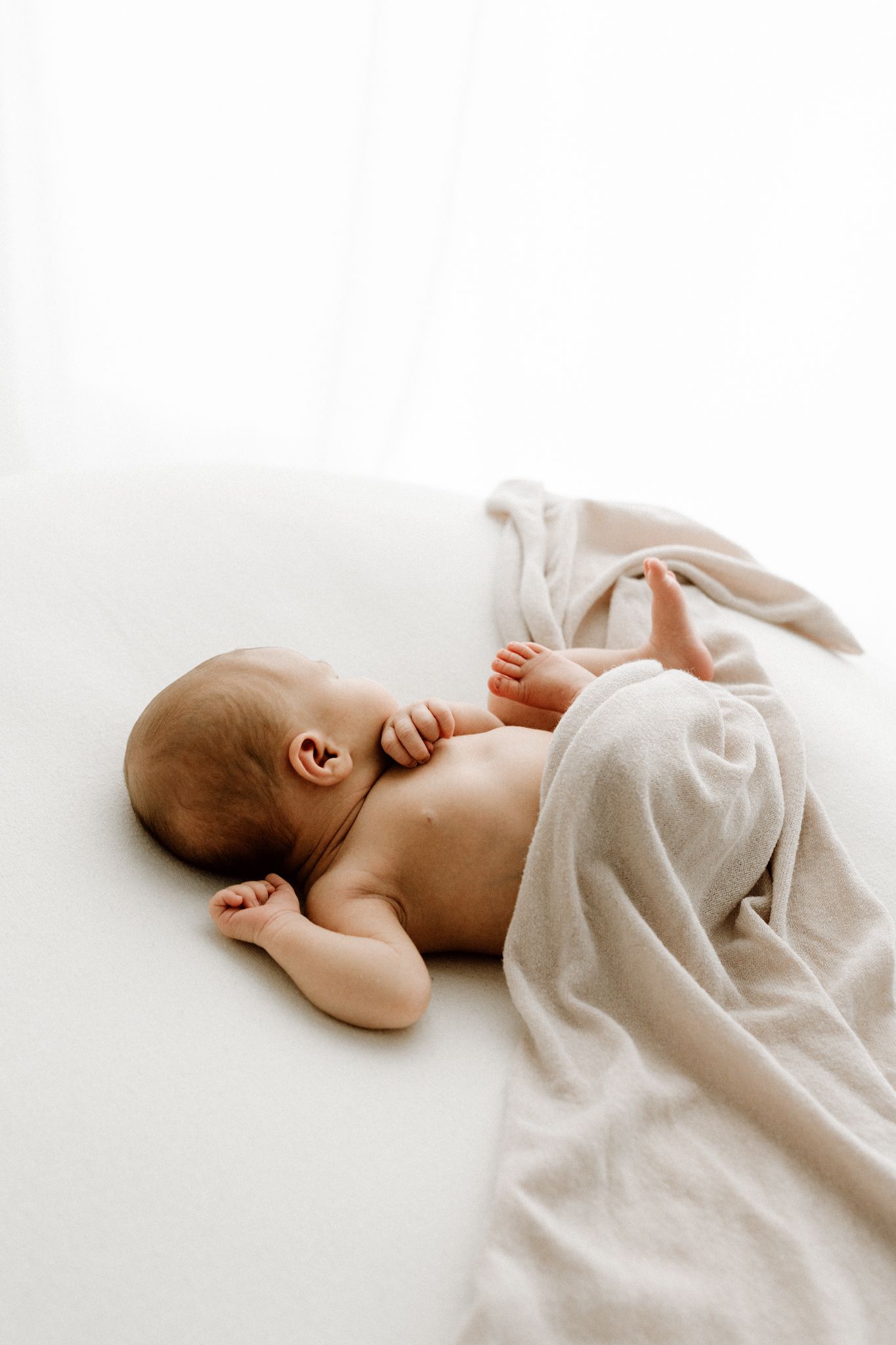 Peaceful newborn baby lying on white surface partially wrapped in beige blanket.