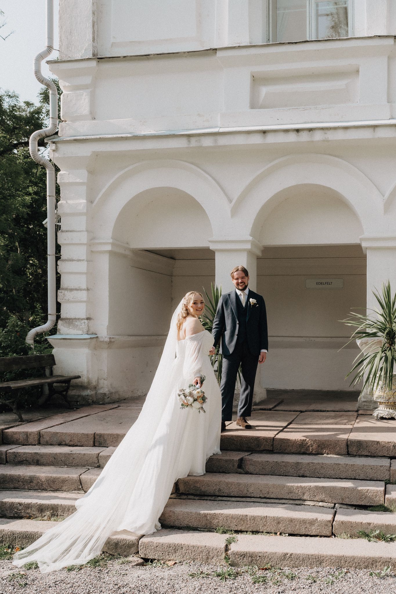 Elegant bride in flowing white gown with groom descending steps of a white classical building.