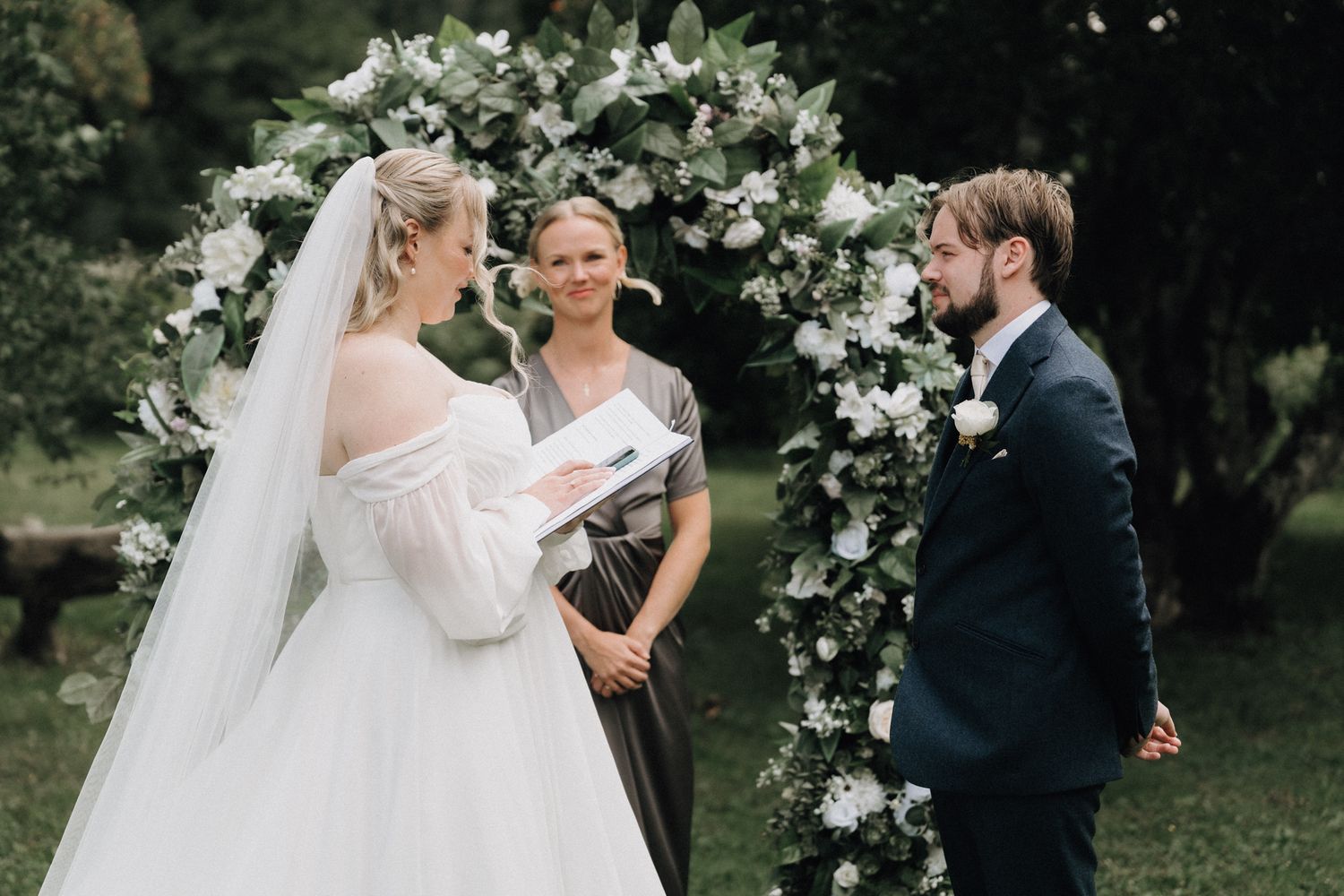 Bride and groom exchange vows at floral arch with officiant during outdoor garden wedding ceremony.