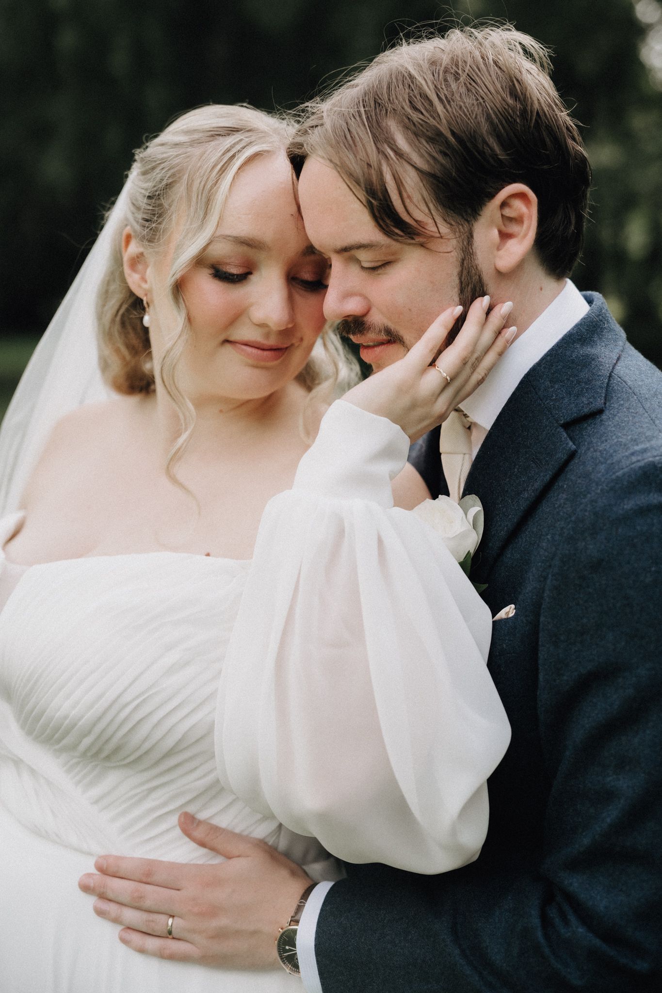 A bride in a white puff-sleeve gown tenderly cups her grooms face as they embrace outdoors.