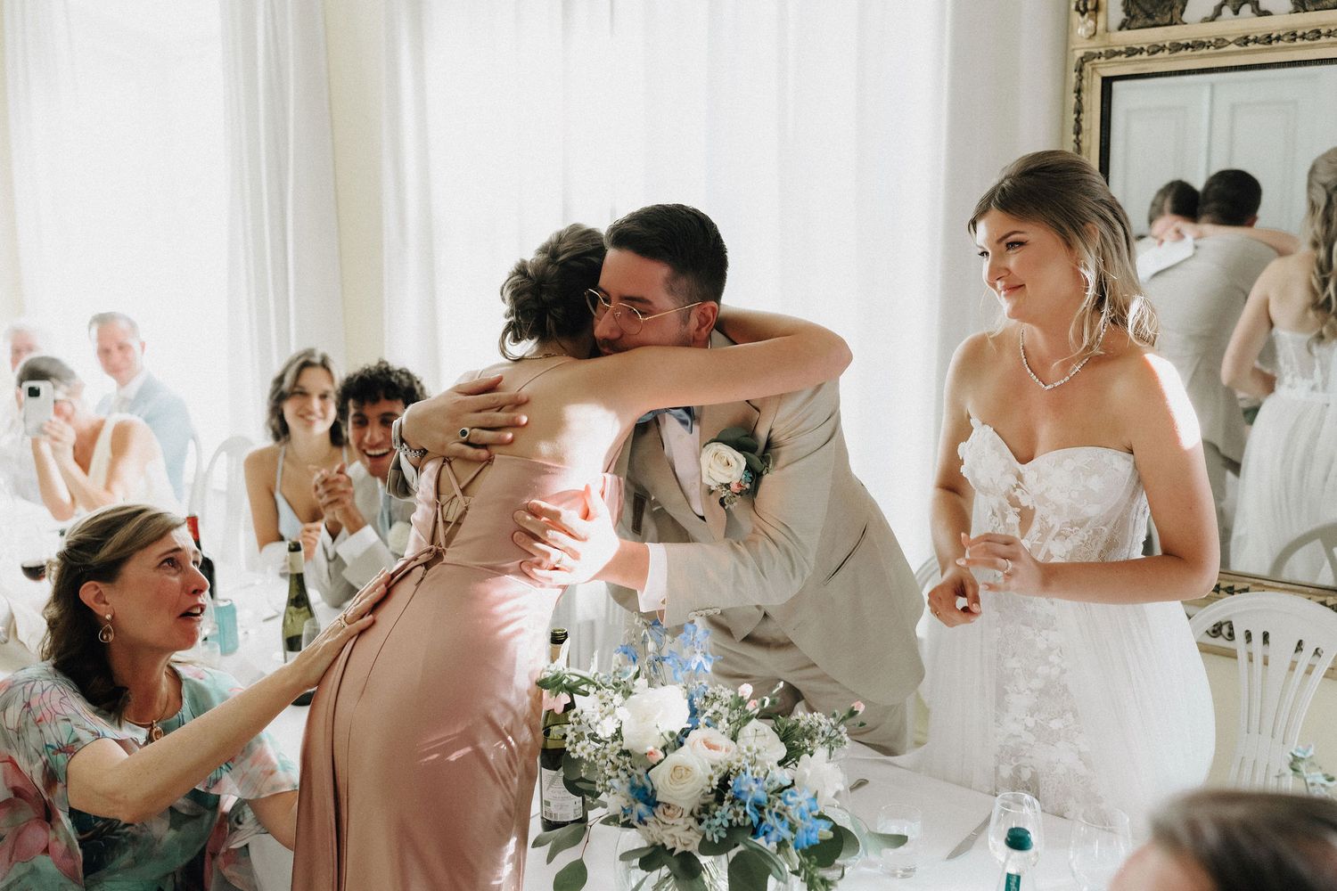 Groom in beige suit embracing bridesmaid in pink dress at wedding reception while bride looks on.
