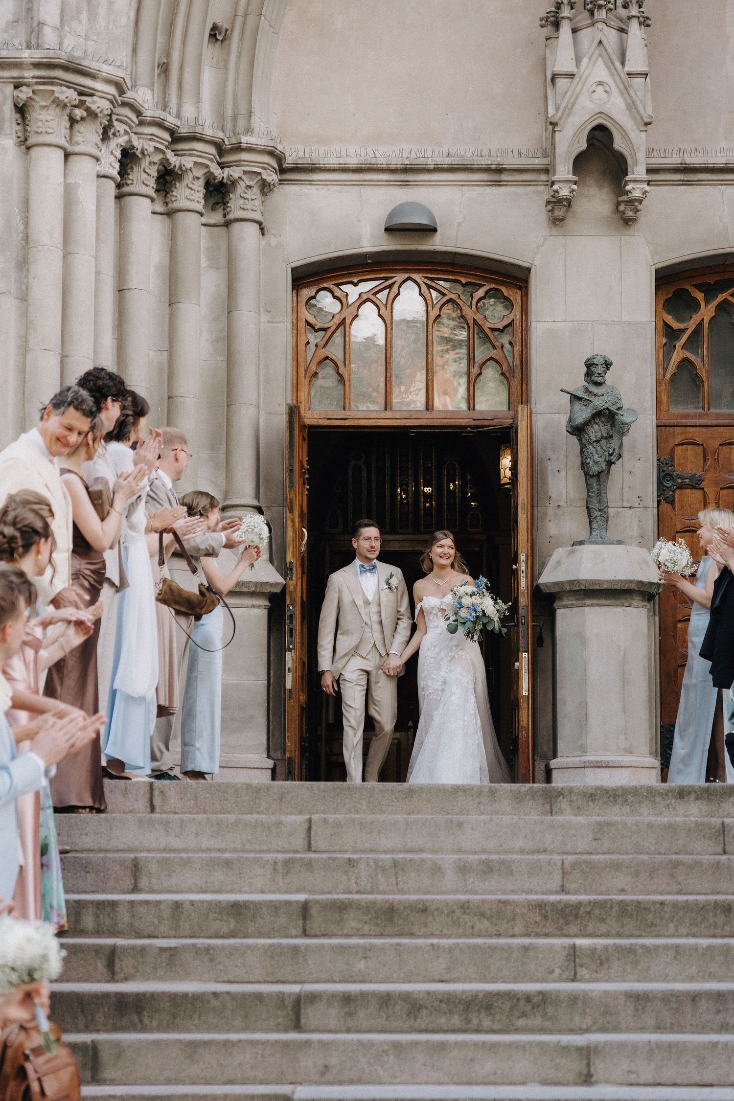 Bride and groom exit a Gothic stone church greeted by cheering wedding guests on the steps.