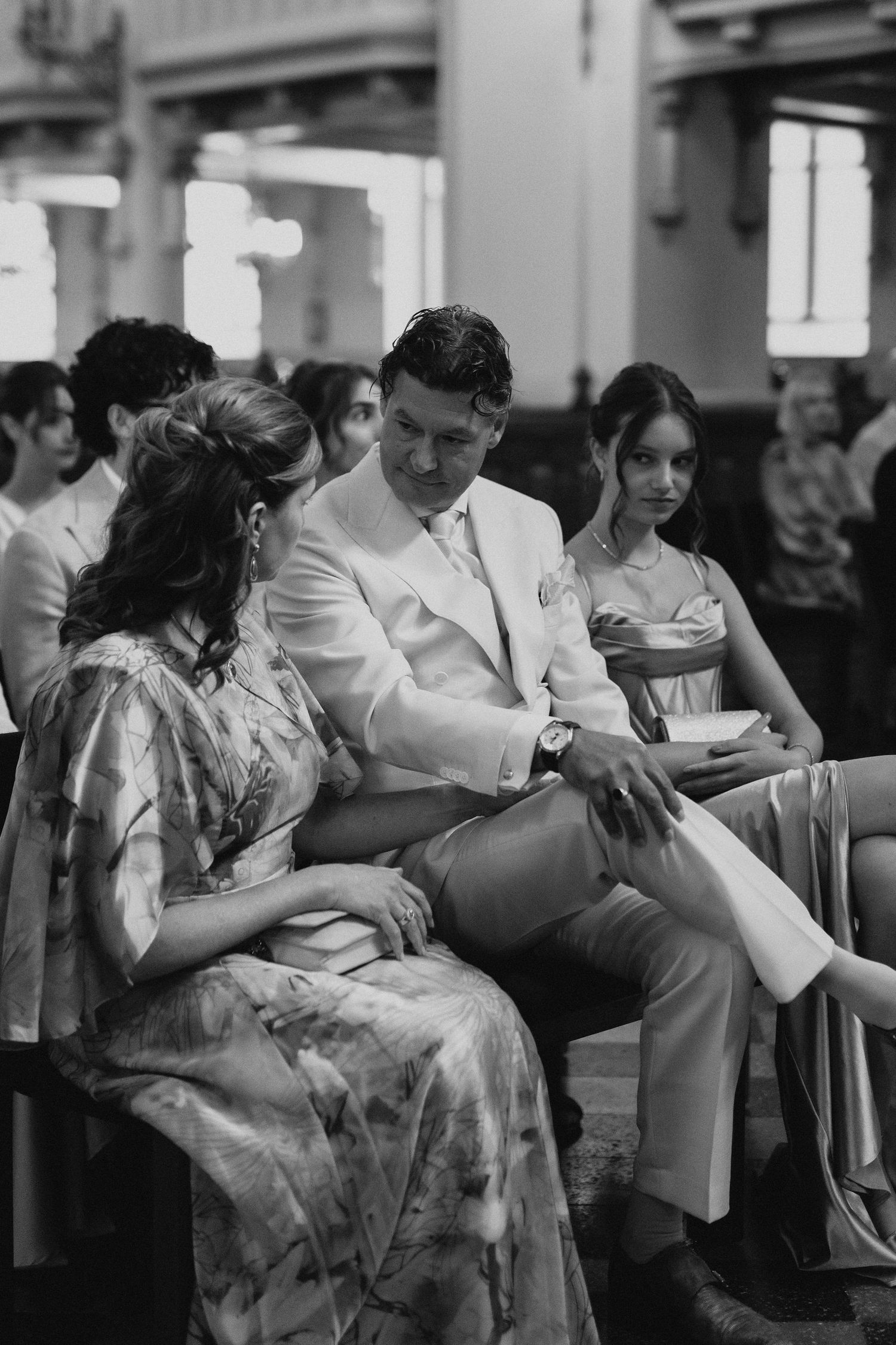 Groom in white suit reviews ceremony program with woman in floral dress in church pew.