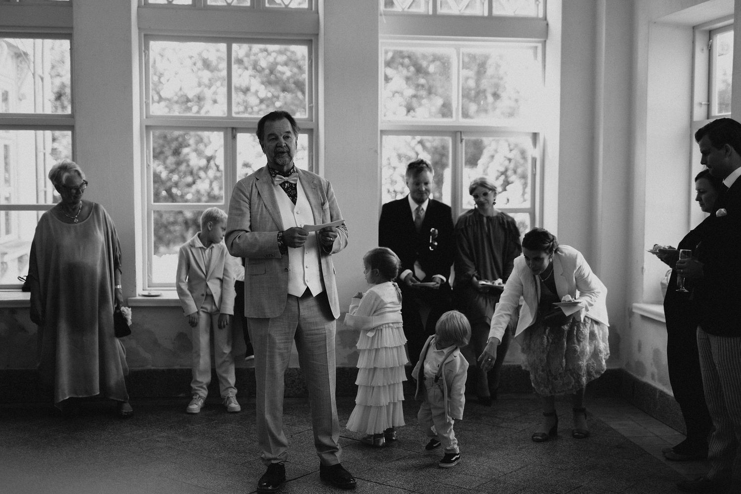 Black and white photo of wedding guests including children gathered in bright windowed venue.