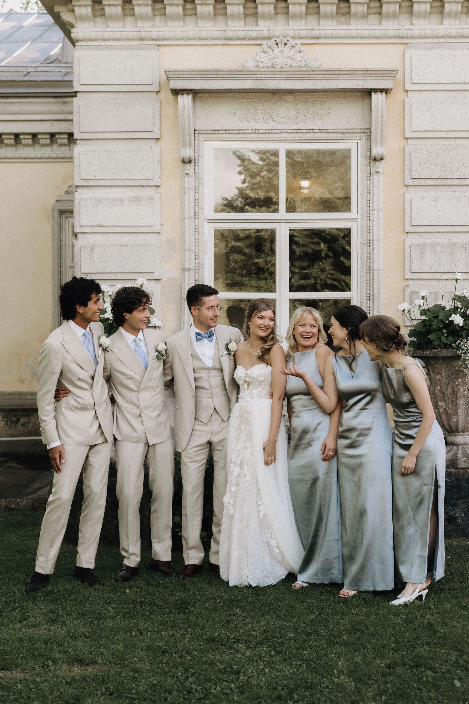 Wedding party laughing together outside classical building, groomsmen in beige suits and bridesmaids in satin dresses.
