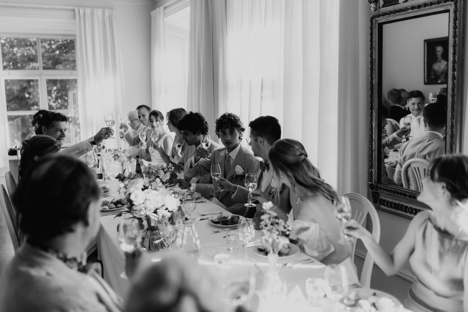 Black and white photo of wedding guests seated at a long decorated reception table with floral arrangements.