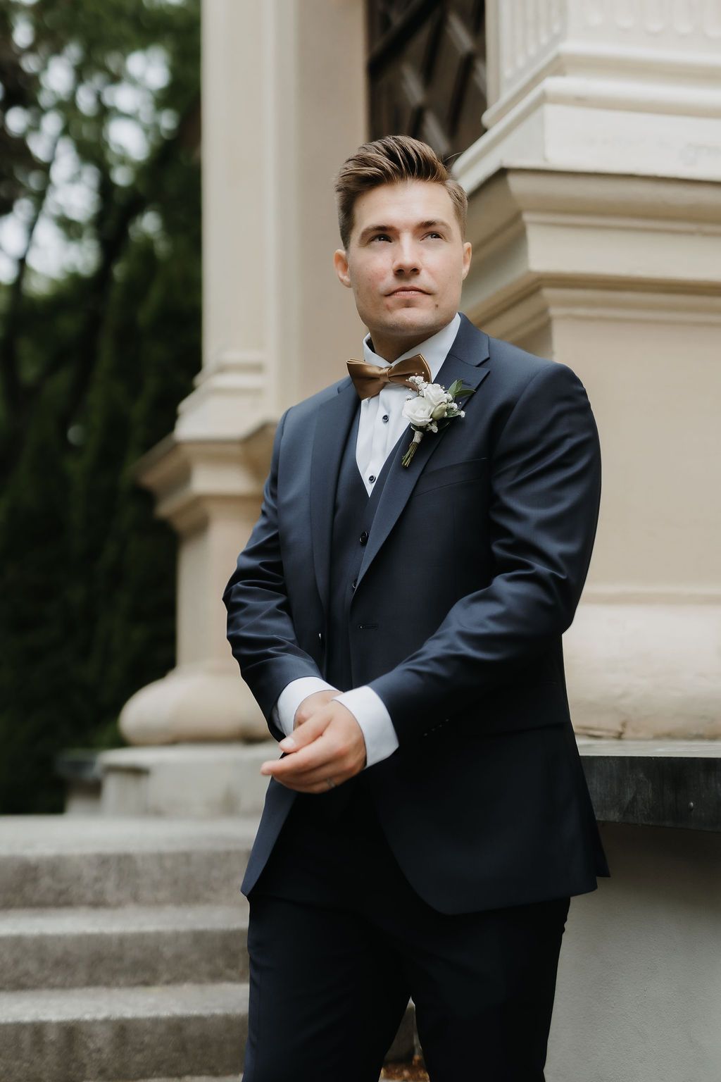 Handsome groom in navy suit and brown bow tie with boutonniere posing at elegant outdoor venue.
