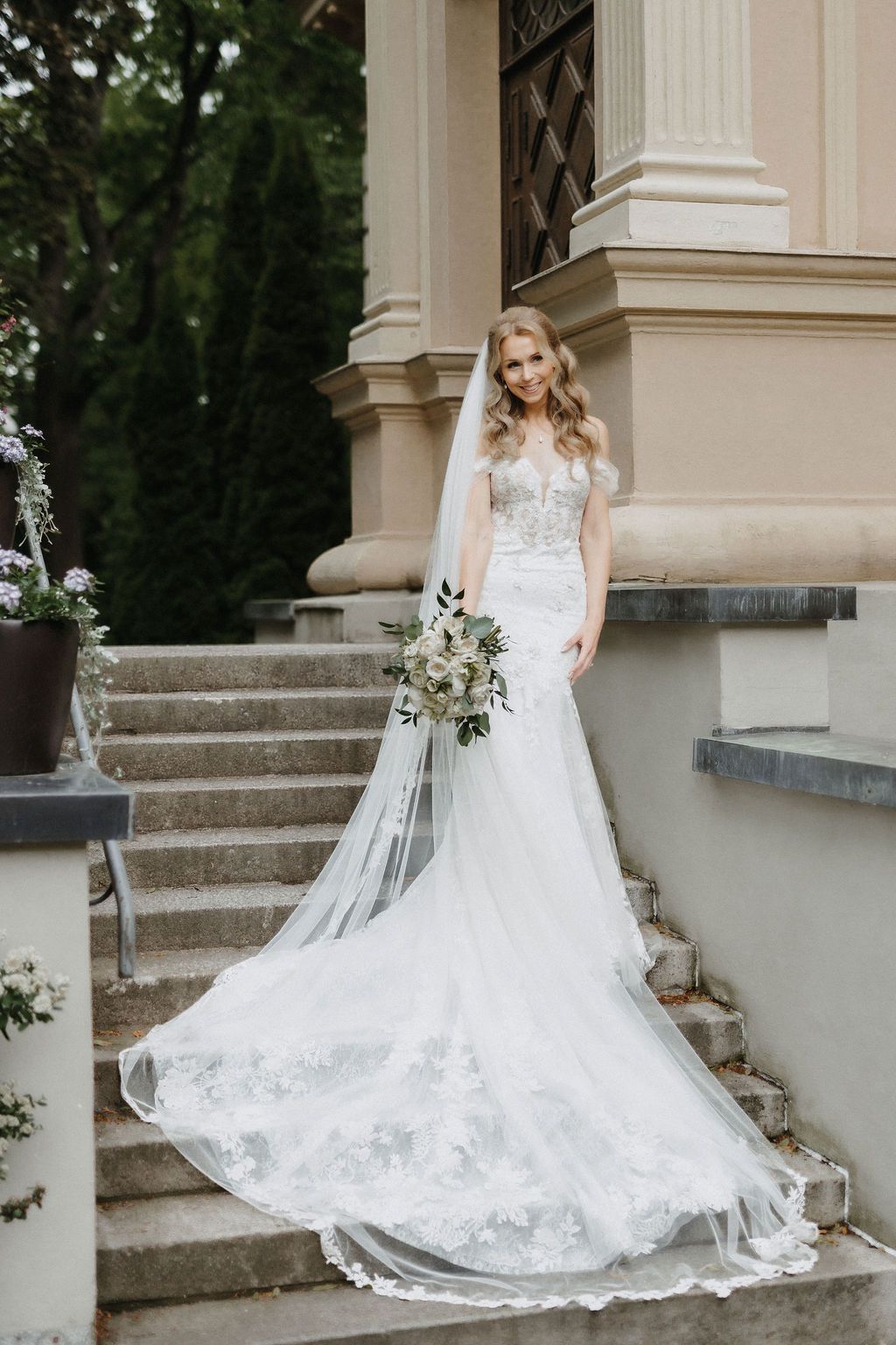 Smiling bride in lace mermaid gown with long veil holds white bouquet on elegant stone steps.