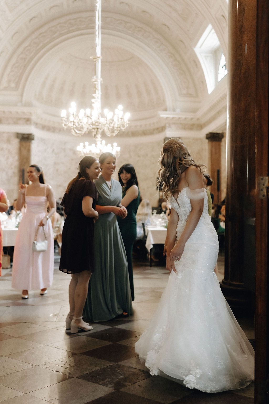 Bride in lace mermaid gown greets guests in elegant chandelier-lit wedding venue hall.
